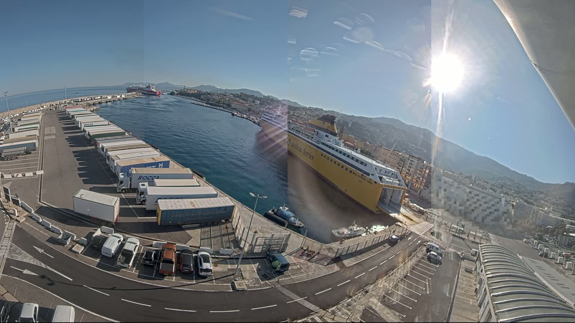 A large yellow ferry is docked at a port on a sunny day, with numerous trucks and cars parked in the surrounding area, and mountains visible in the background.