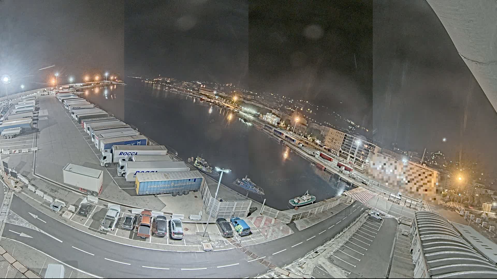 A nighttime, high-angle view shows a harbor with numerous parked semi-trailer trucks, several cars, and a few small boats on the water.