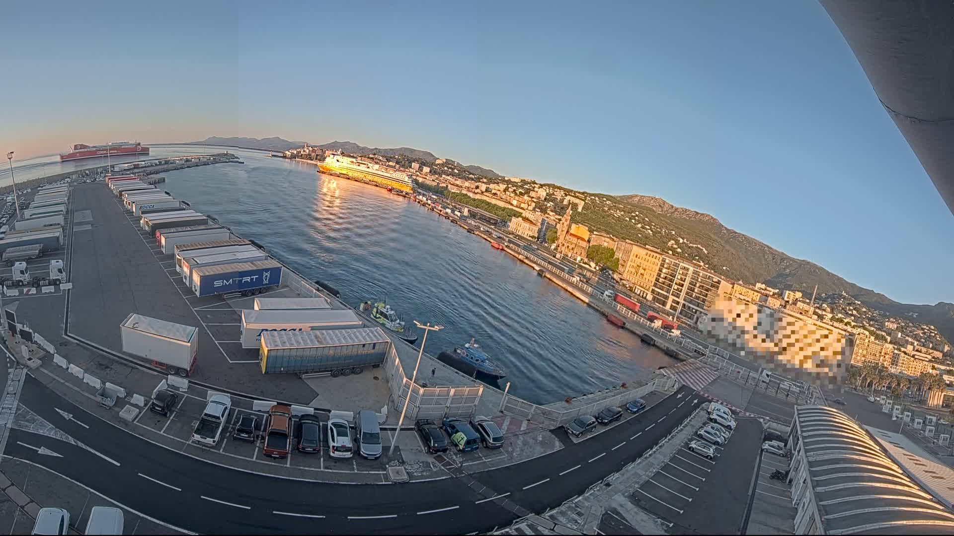 A harbor filled with docked ferries and numerous semi-trailer trucks parked at a terminal, under a clear, sunny sky, with mountains in the background.