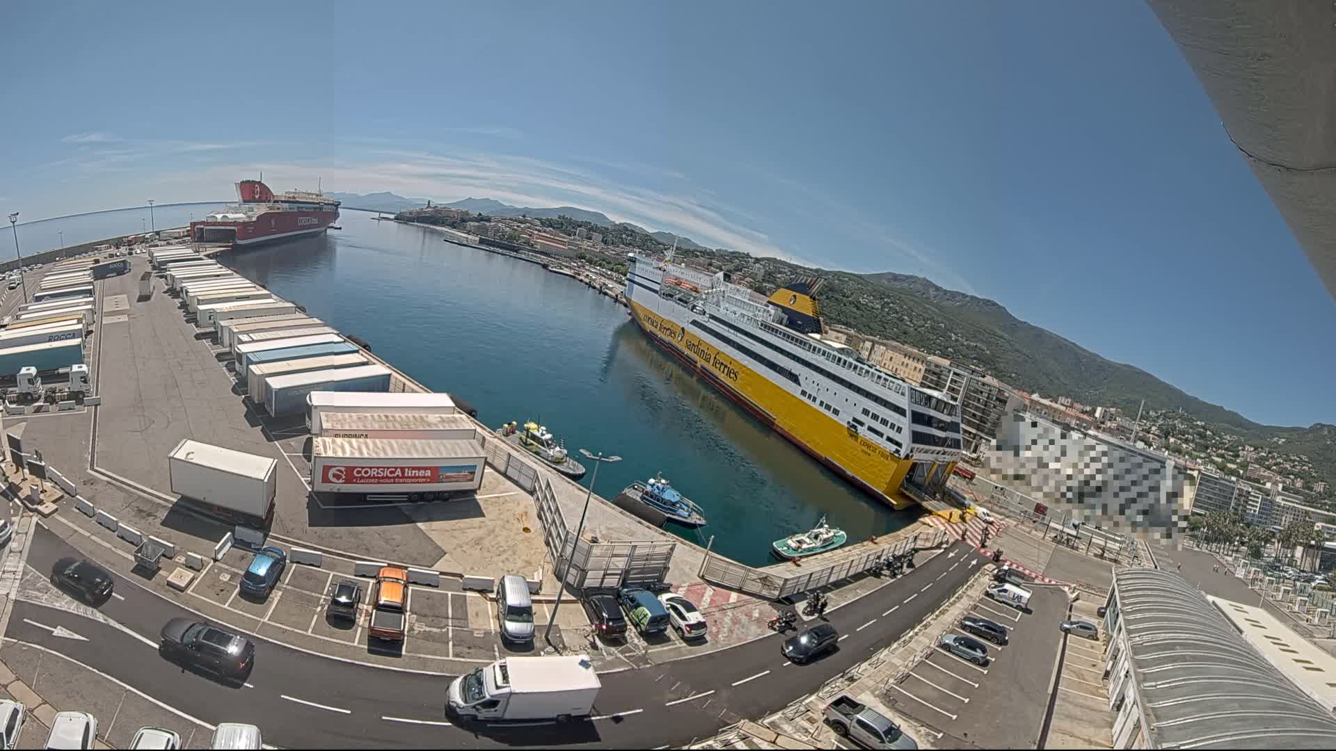 A sunny day at a port shows two large ferries docked, numerous cargo trucks and cars in a parking lot, and smaller boats in the water, all surrounded by buildings and mountains in the distance.