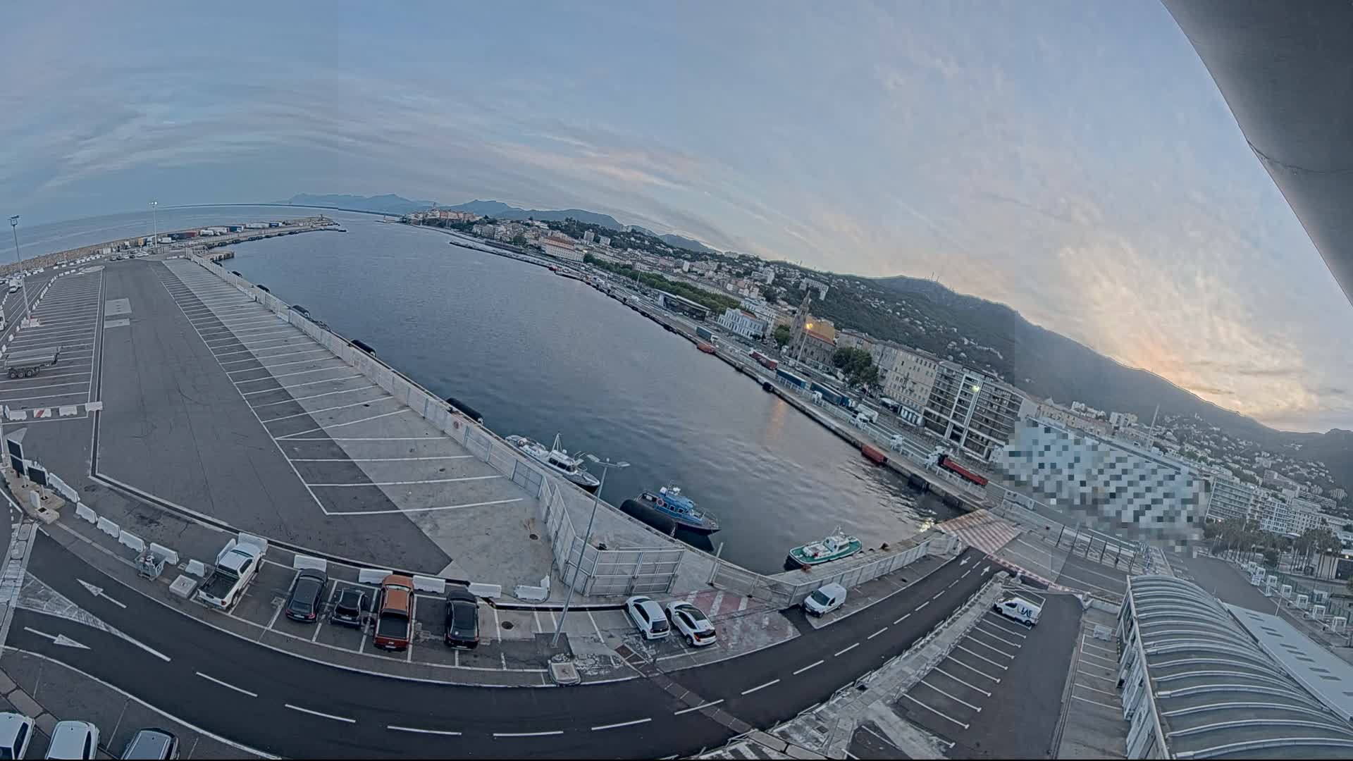 An expansive harbor with a large empty parking lot, several small boats, and a city nestled against a mountain range under a mostly clear, twilight sky.