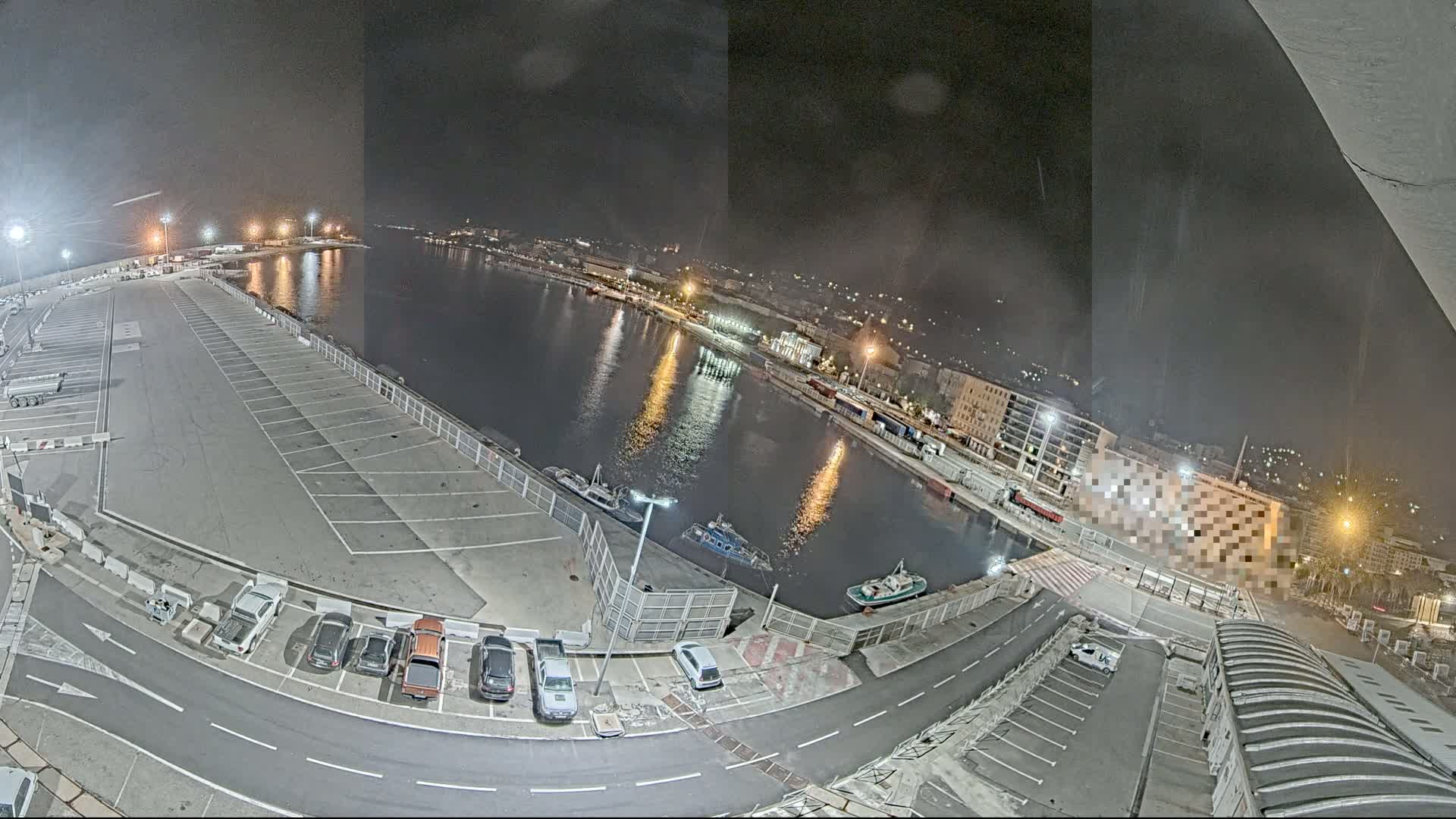 A nighttime view of a harbor with a mostly empty parking lot, several cars, and a few small boats on the water.