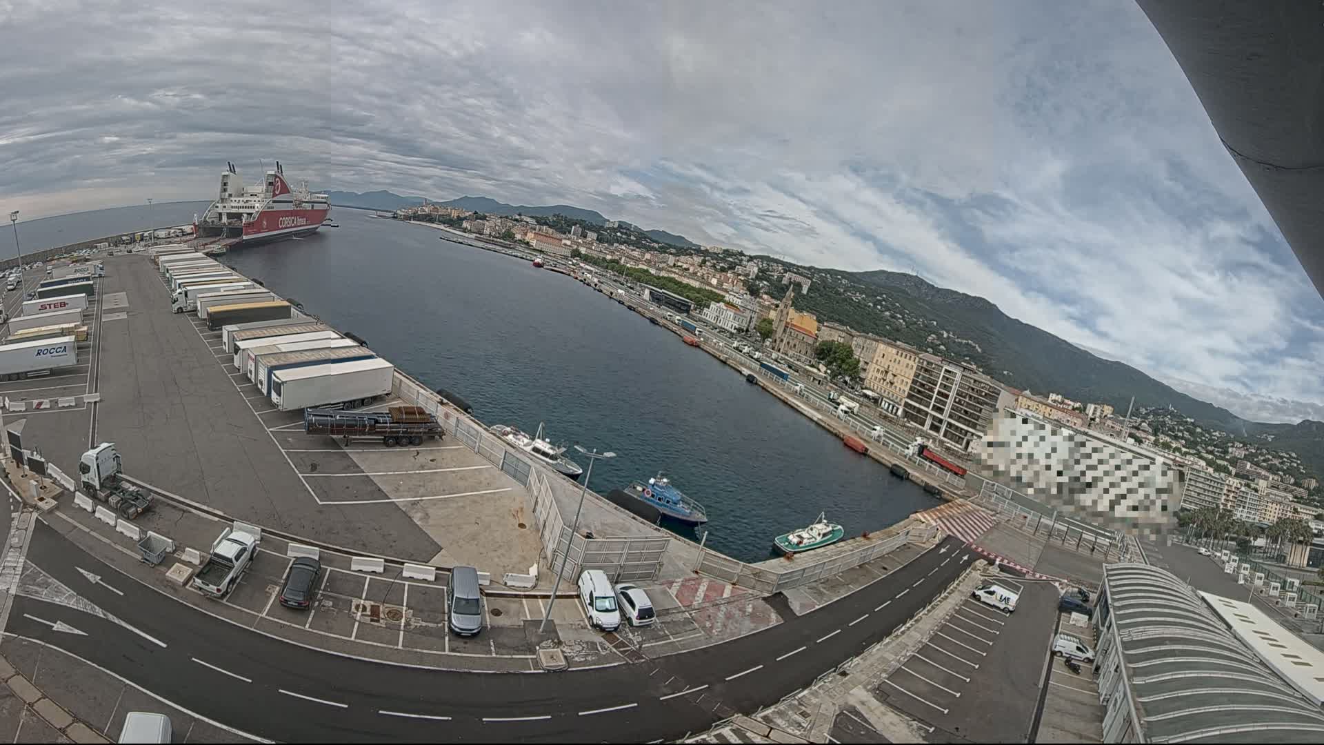 A ferry is docked at a harbor in a city with overcast skies, surrounded by parked trucks, cars, and smaller boats.