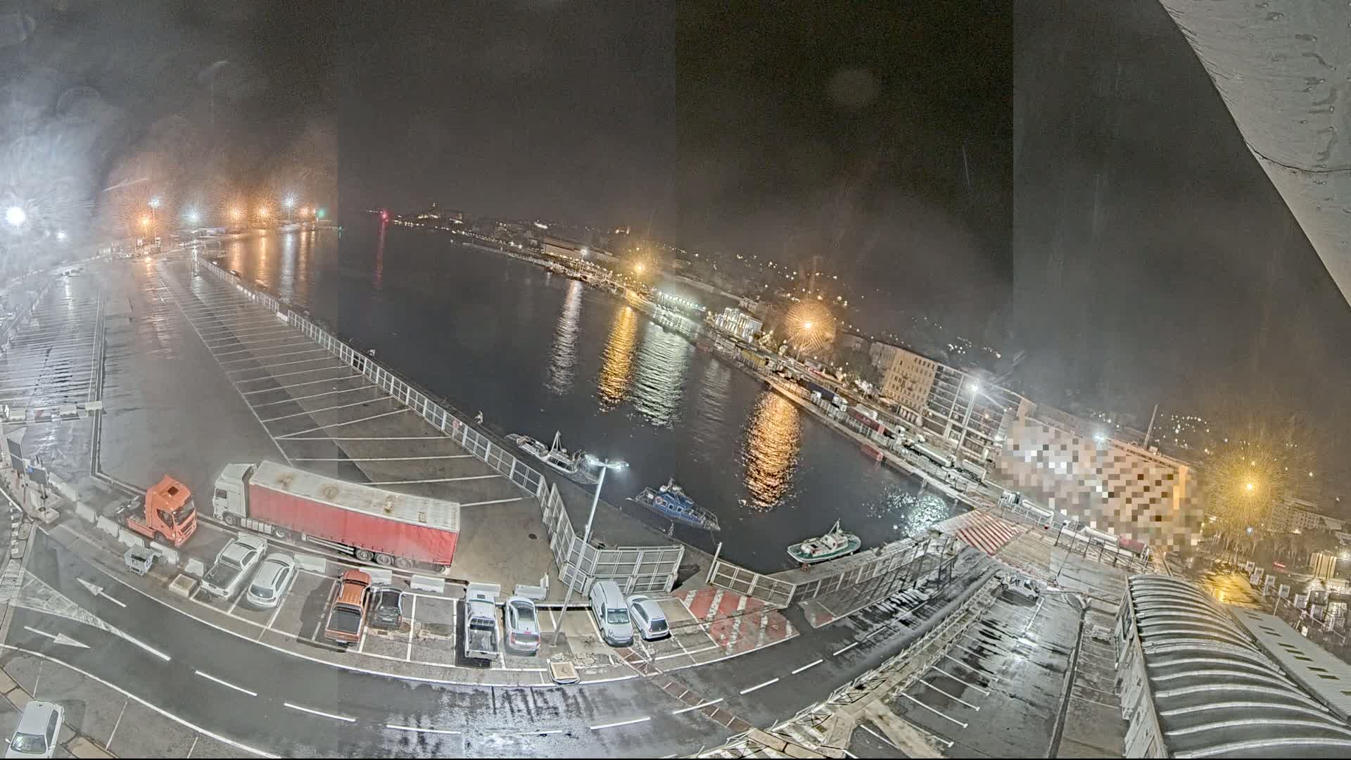 A nighttime view of a harbor with a wet parking lot filled with cars and trucks, several boats on the water, and buildings illuminated with lights along the shoreline under slightly misty conditions.