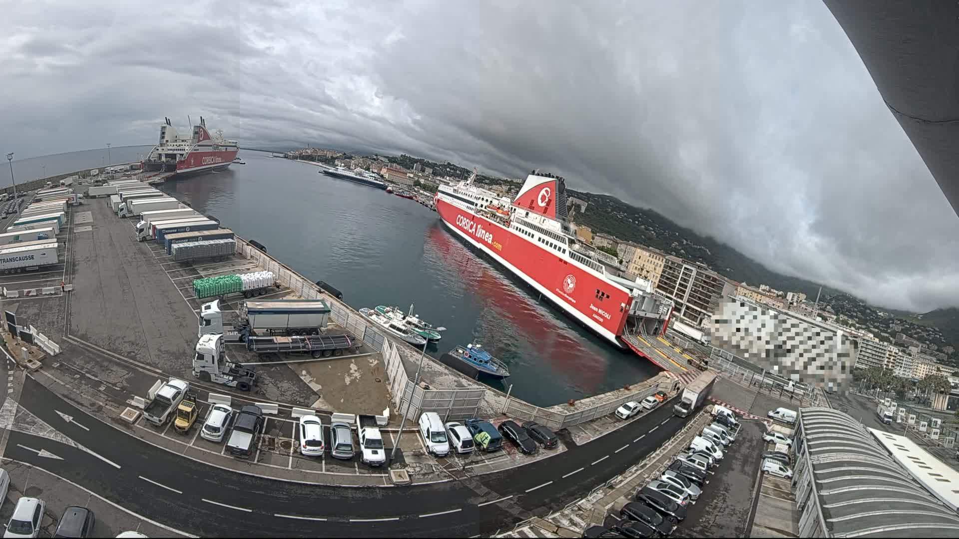 A large red ferry is docked at a port under a cloudy sky, with numerous cars and trucks parked in the surrounding area.