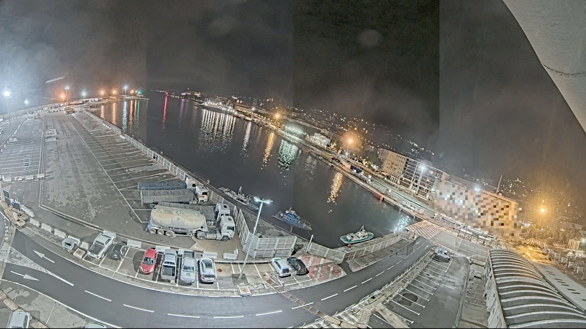 A nighttime aerial view of a harbor with a large empty parking lot, several vehicles, and boats on the water.