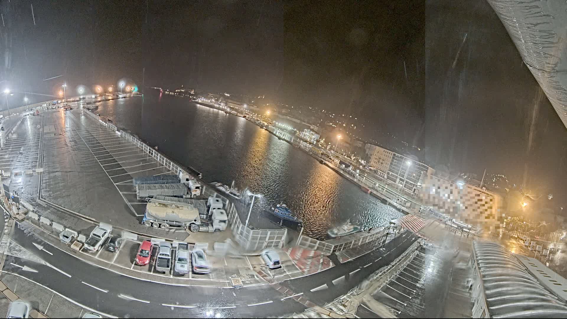 A nighttime, rain-soaked harbor scene shows a wet parking lot filled with cars and trucks next to a calm body of water with docked boats and buildings along the shoreline.