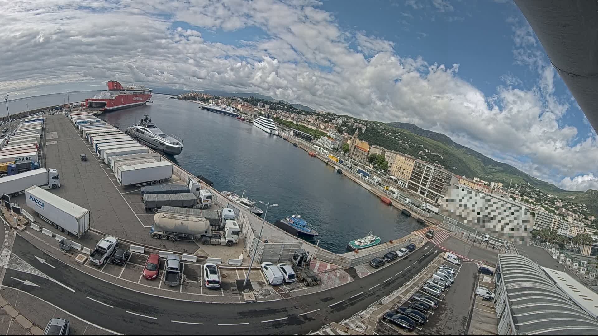 A harbor scene under partly cloudy skies shows a large red ferry, a yacht, numerous trucks and cars parked in lots, and smaller vessels in the water near a city at the base of mountains.