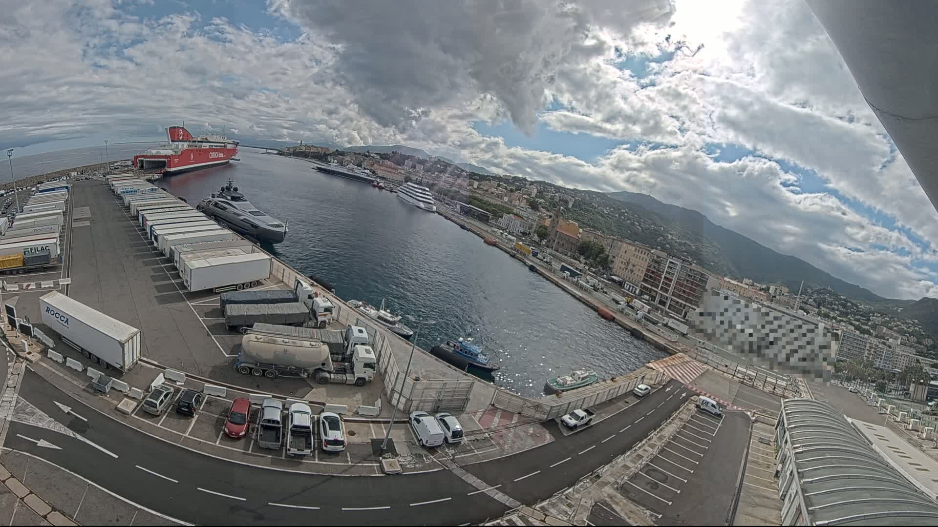 A harbor with a large red ferry, a yacht, and numerous trucks and cars parked, under a partly cloudy sky.