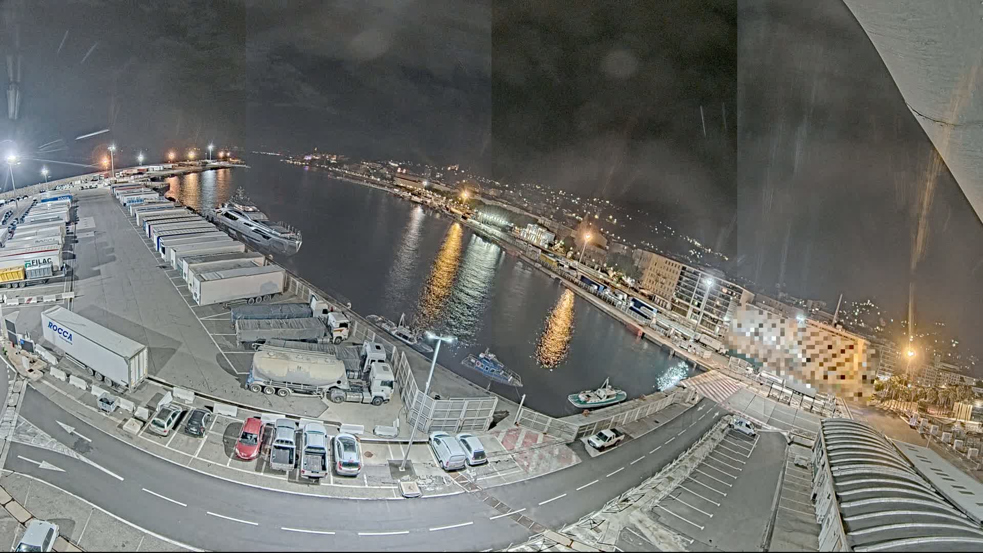 A nighttime, high-angle view of a harbor with numerous parked trucks and cars, boats in the water, and city lights in the background under a partly cloudy sky.