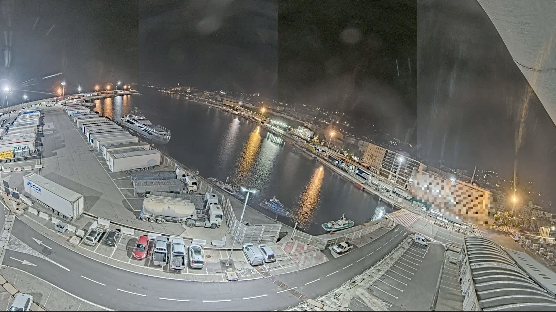 A nighttime aerial view of a harbor, showing a parking lot filled with trucks and cars, a calm body of water with several boats, and city lights in the background under a lightly drizzling sky.