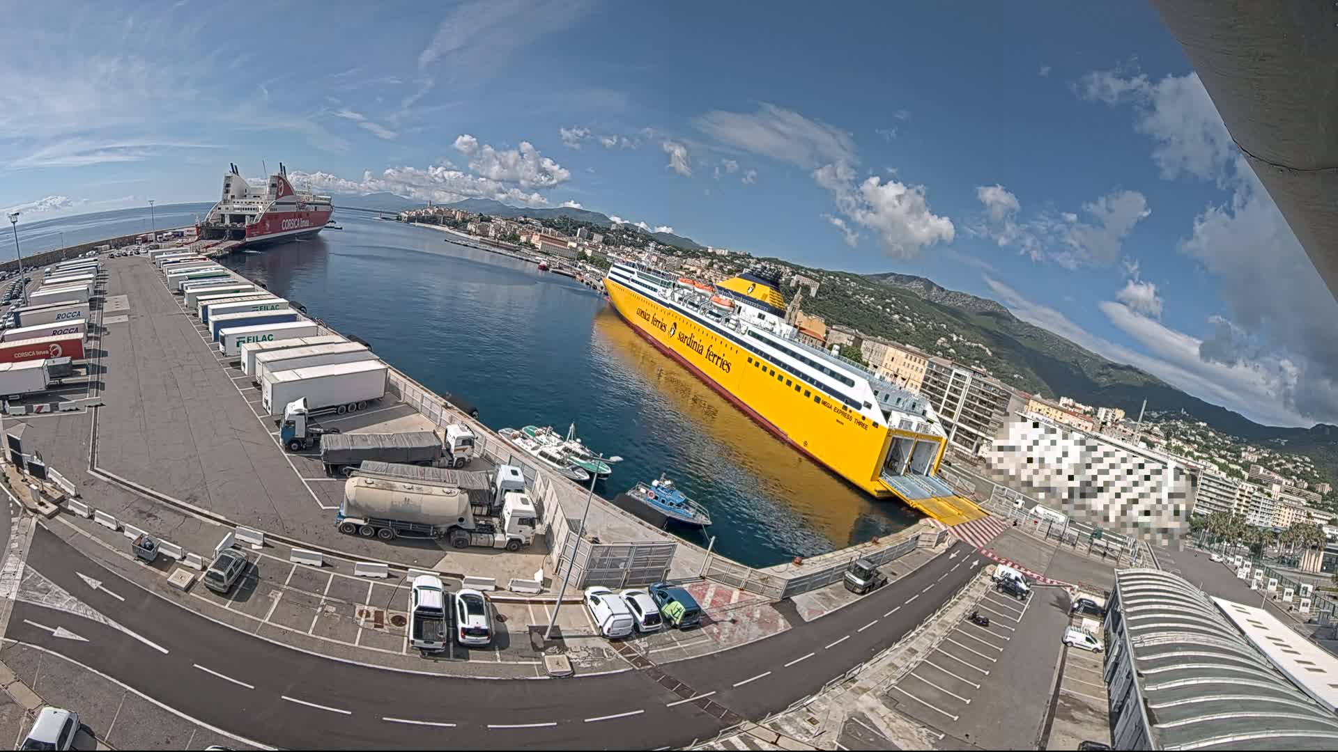 A yellow and white ferry is docked at a port with several other vehicles and trailers parked nearby, under a mostly sunny sky.