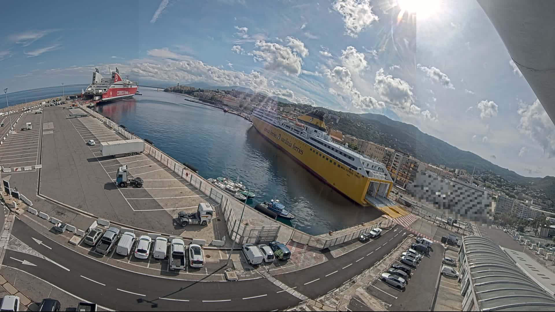 A sunny day at a port shows two large ferries docked, numerous cars parked in lots, and a city and mountains in the background.