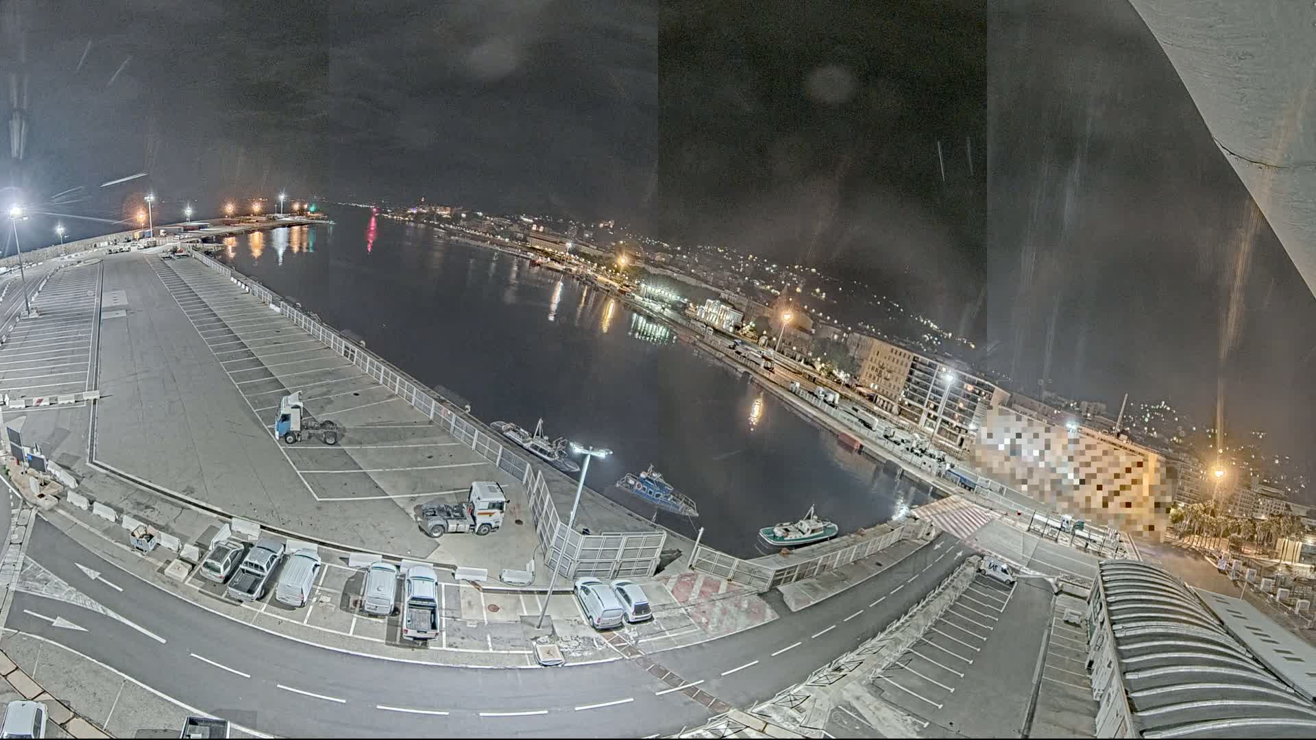 A nighttime, high-angle view of a harbor with a large empty parking lot, several vehicles, and boats moored alongside a waterfront lined with buildings.
