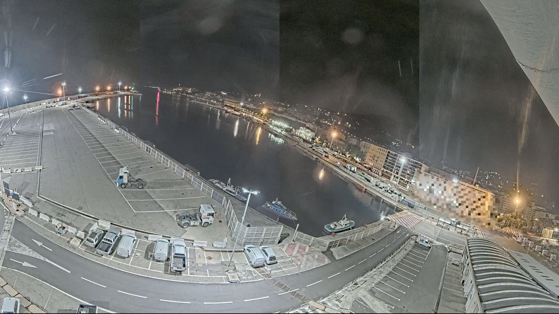 A nighttime aerial view of a harbor with a large empty parking lot, several vehicles, and boats moored along a calm body of water next to a city.