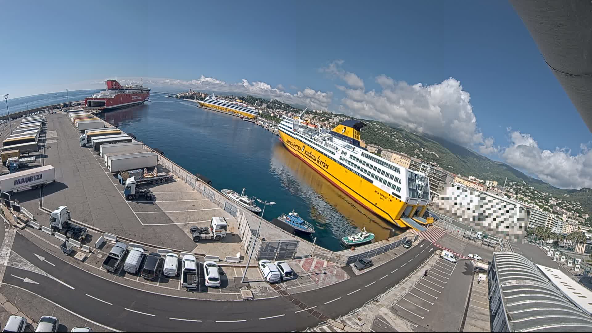 A harbor on a sunny day with partly cloudy skies shows several ferries docked, along with numerous trucks and cars parked in a lot.