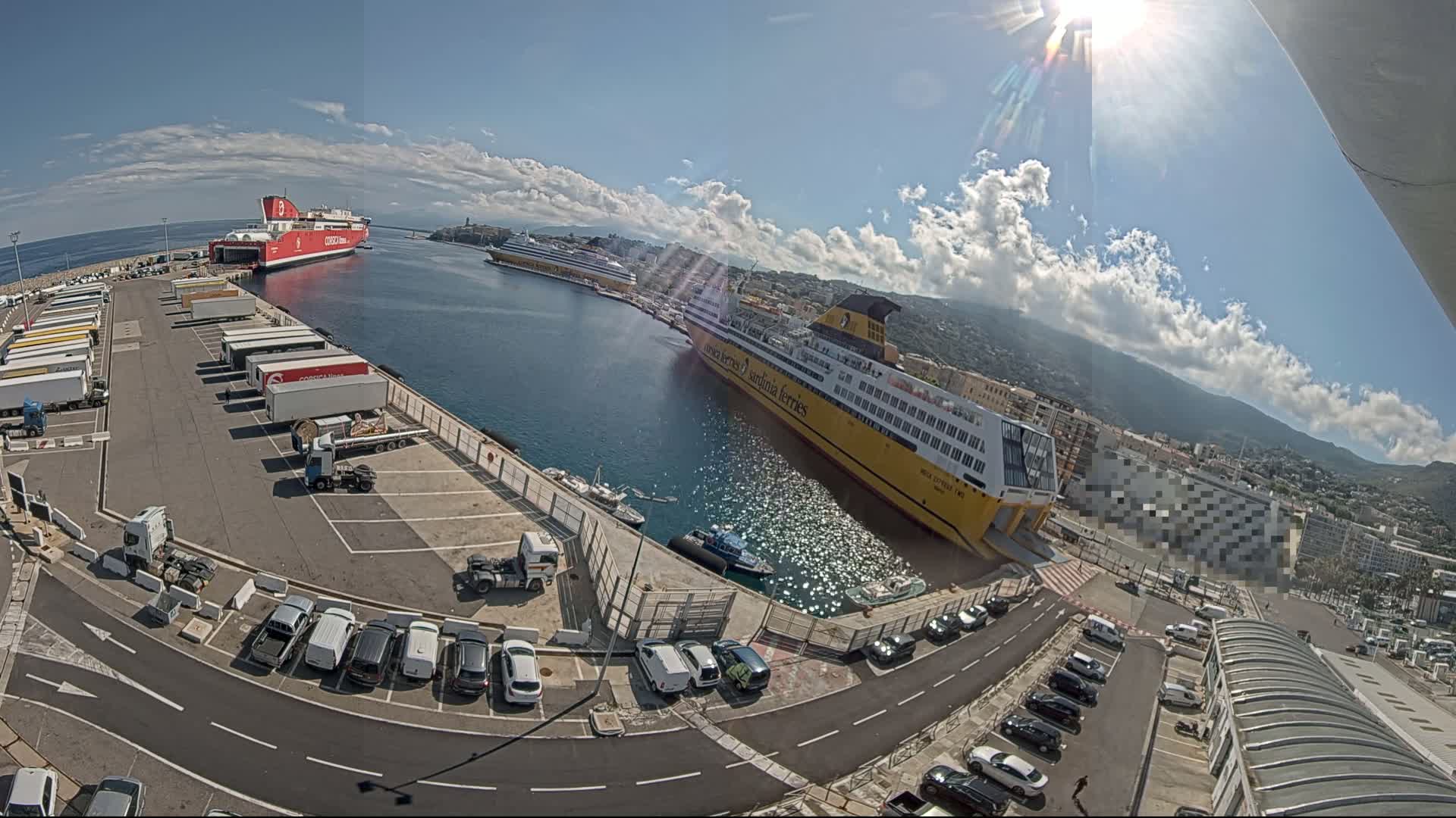 A sunny day at a harbor shows two large ferries docked, with numerous cars and trucks parked in a lot nearby.