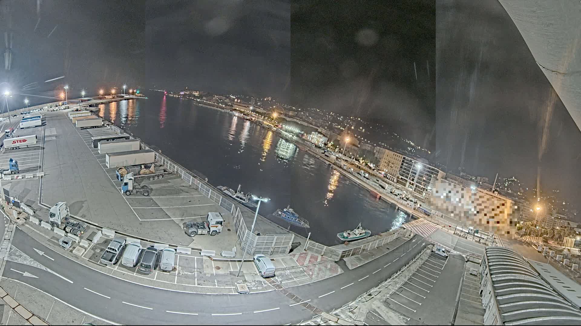 A nighttime, high-angle view shows a harbor with several docked boats, a parking lot filled with trucks and cars, and a city in the background under a slightly overcast sky.