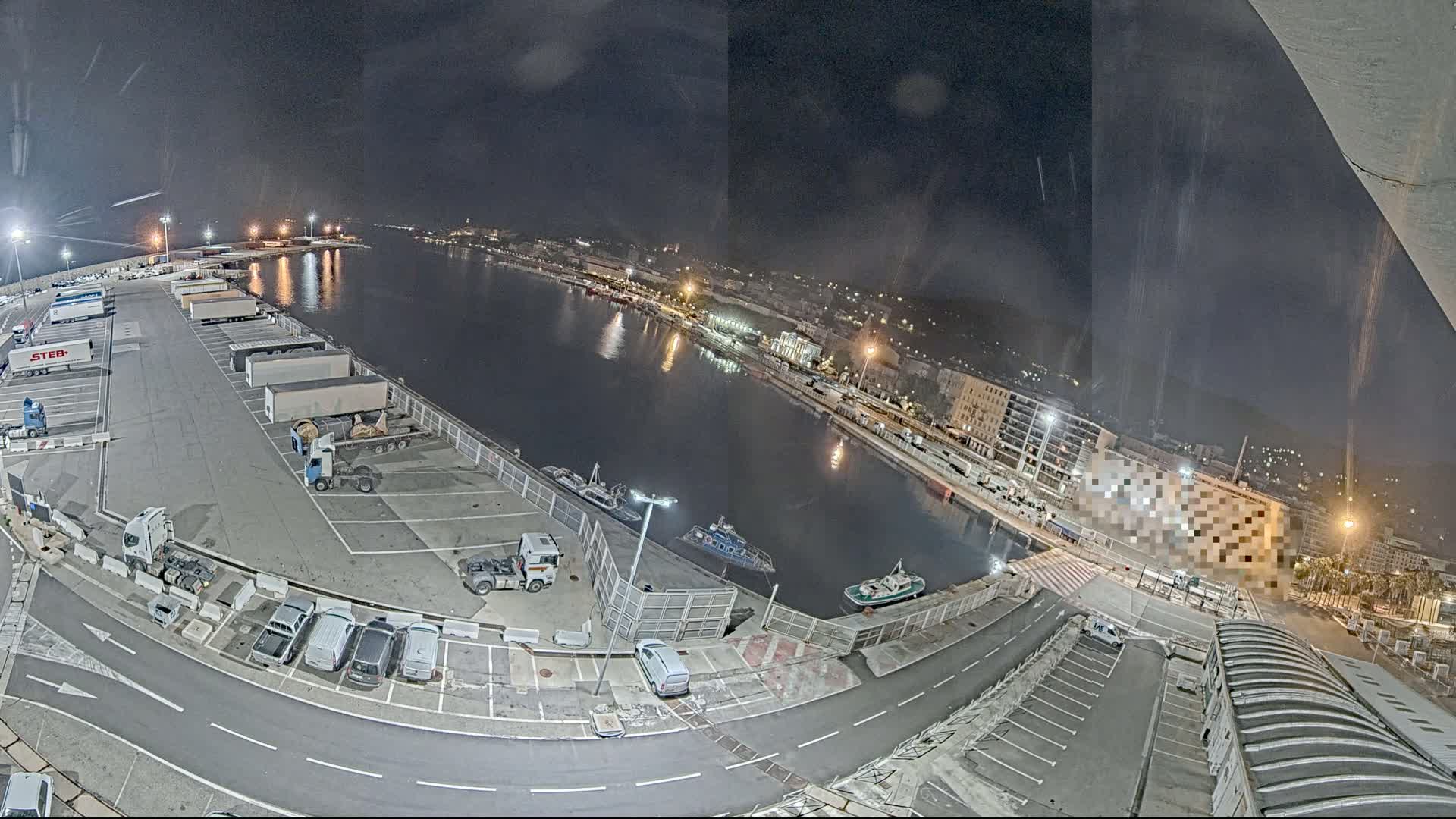 A nighttime, high-angle view of a harbor with a parking lot full of trucks and cars, a body of water with several boats, and city buildings in the background.