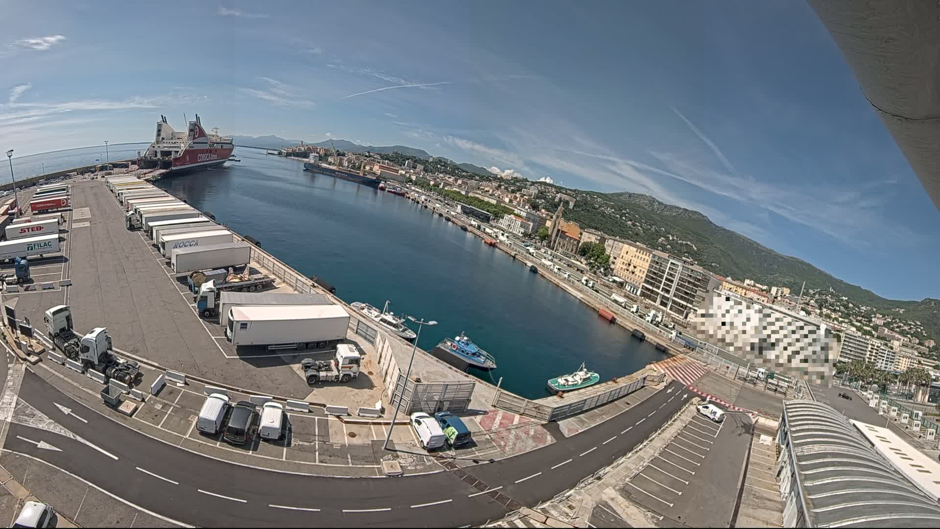 A ferry is docked at a harbor on a sunny day, with numerous trucks and cars parked nearby, and smaller boats in the water.