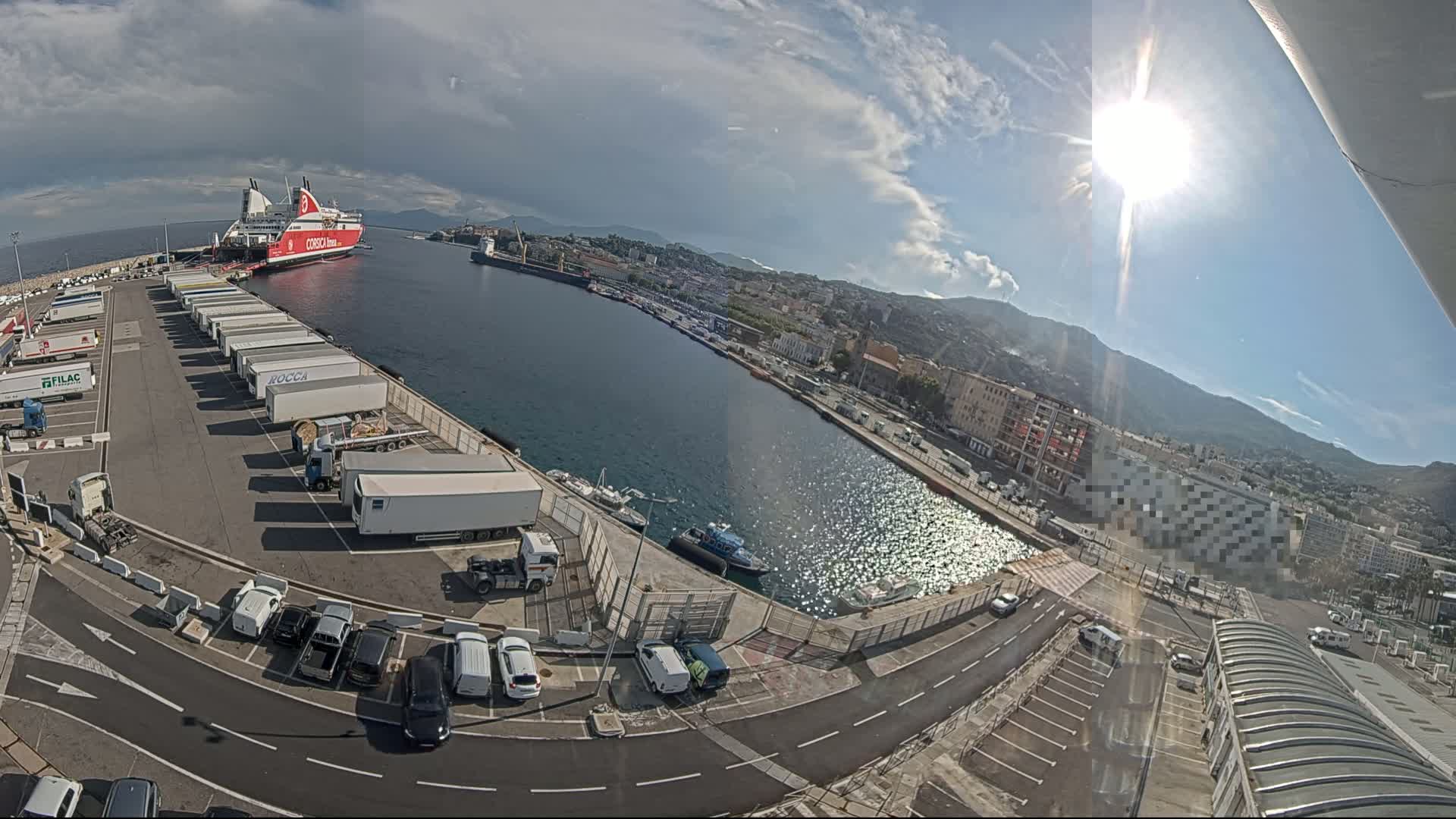 A ferry is docked at a harbor on a sunny day with partly cloudy skies, next to a large parking lot filled with semi-trailer trucks and cars.