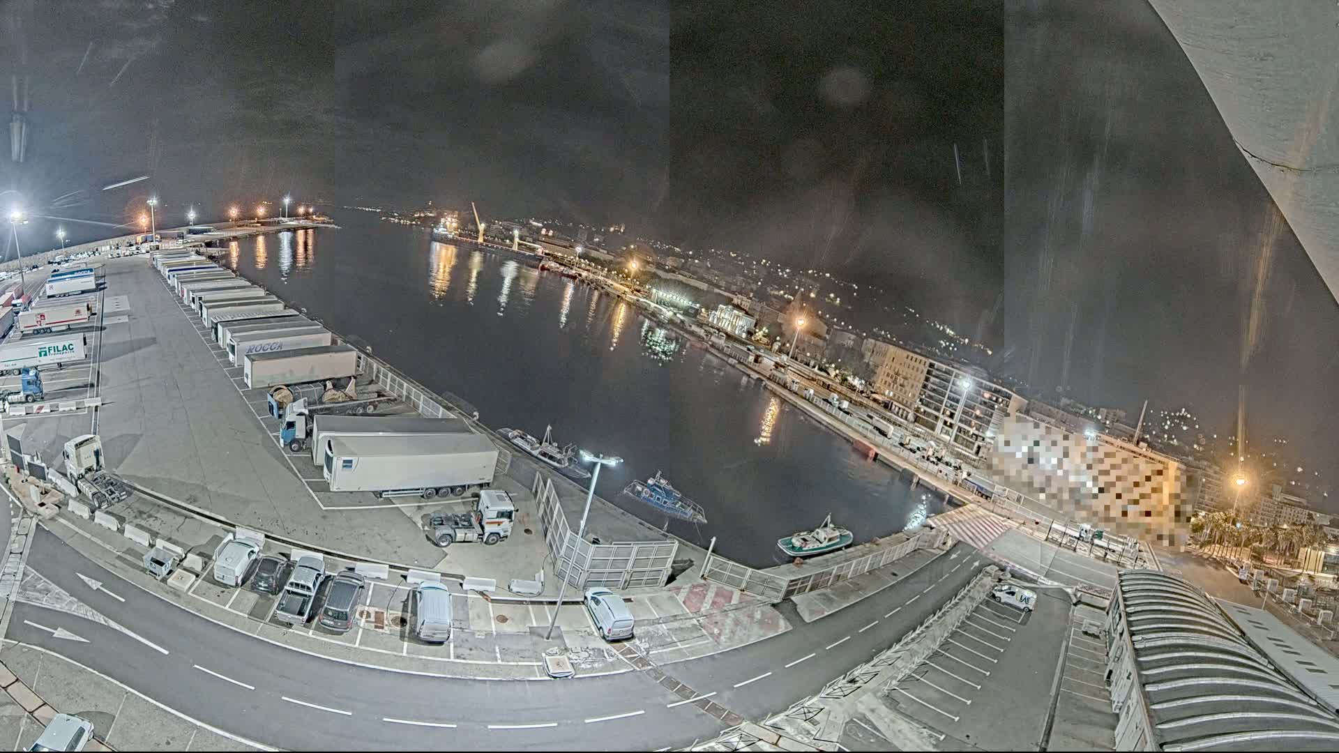 A nighttime, high-angle view of a harbor shows numerous trucks parked in a lot next to a calm body of water, with several small boats and buildings visible in the distance.