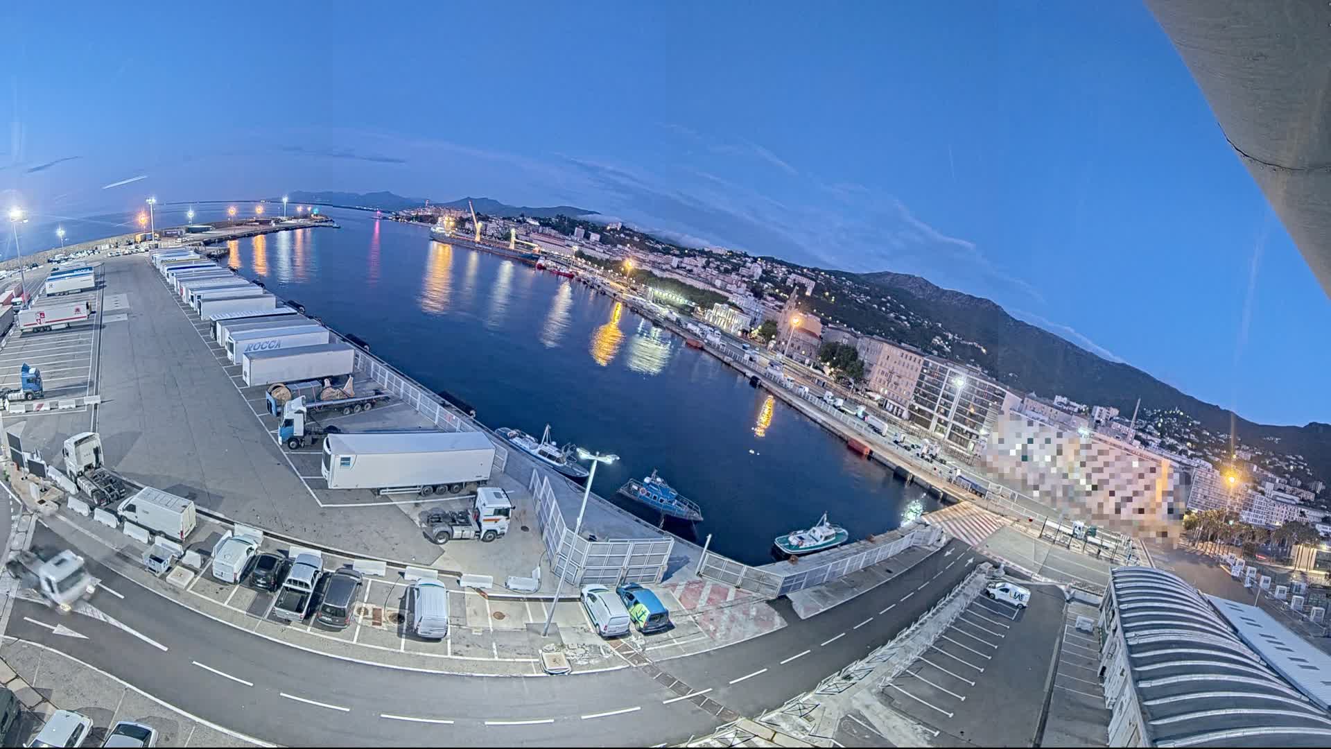 A high-angle, twilight view of a harbor with numerous trucks parked in a lot, several small boats in the water, and a city rising on a hillside in the background.