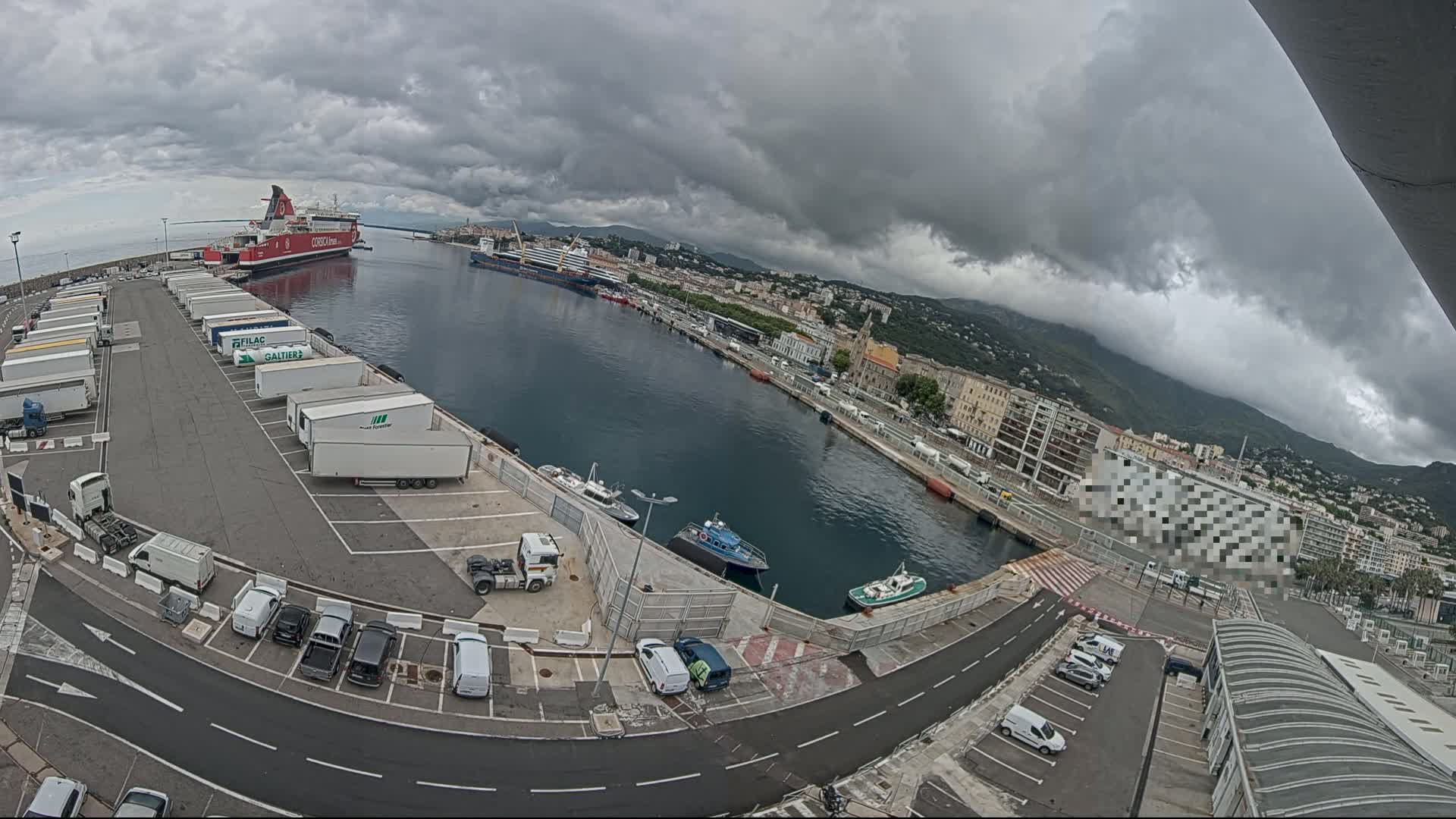 A ferry and other ships are docked at a harbor under an overcast sky, with numerous trucks and cars parked in the surrounding lots.