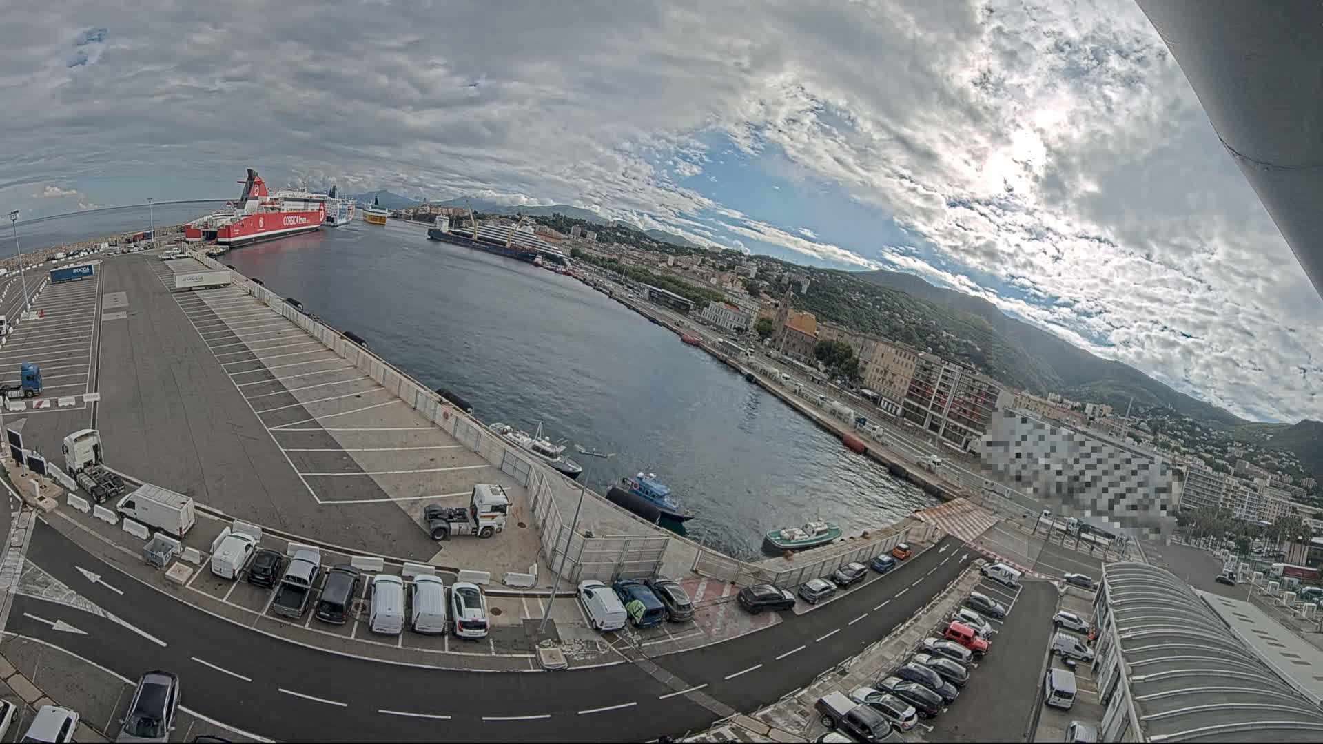A partly cloudy day shows a harbor with several docked ships, a large empty parking lot filled with cars, and a city in the background.