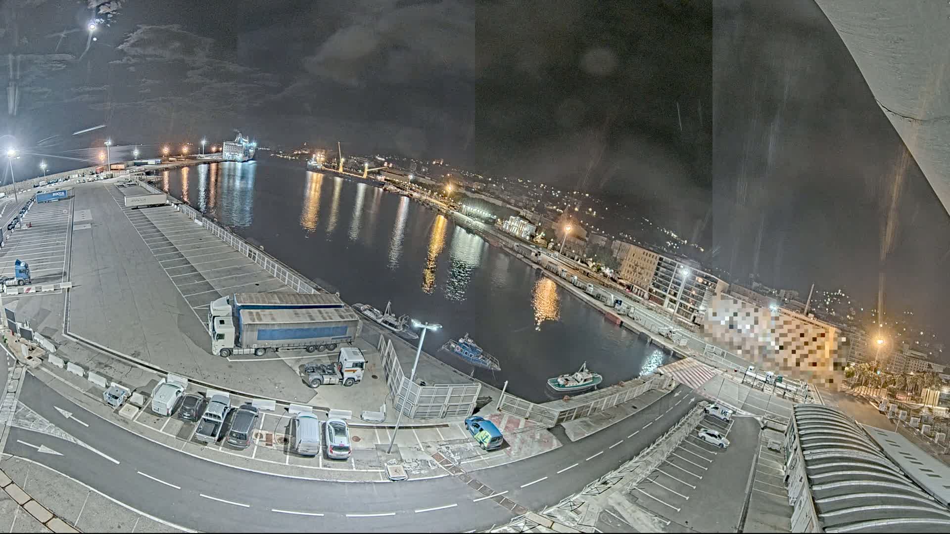 A nighttime view of a harbor with parked cars and trucks, a few small boats, and a larger ship in the water under a cloudy sky.