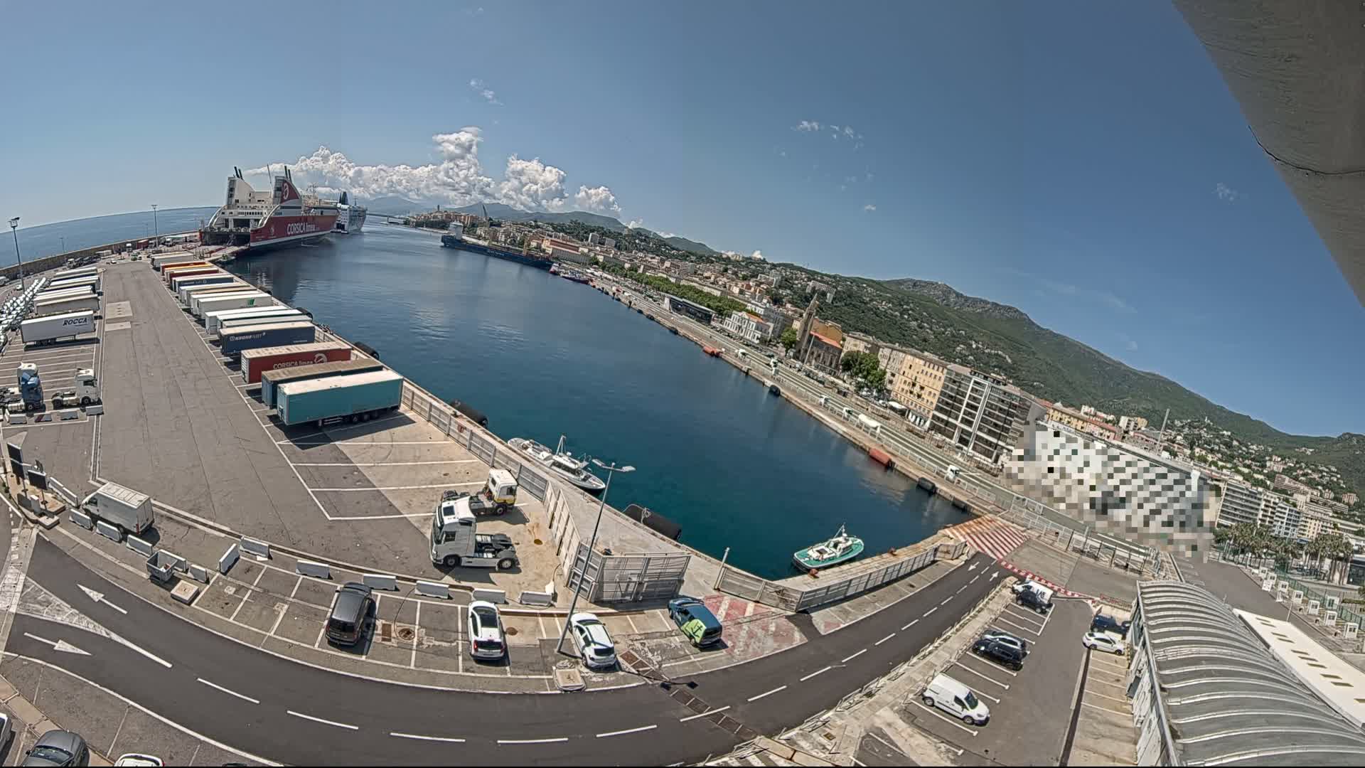 A large ferry is docked at a harbor on a sunny day, with numerous trucks and cars parked in the surrounding lots, and buildings and mountains visible in the background.