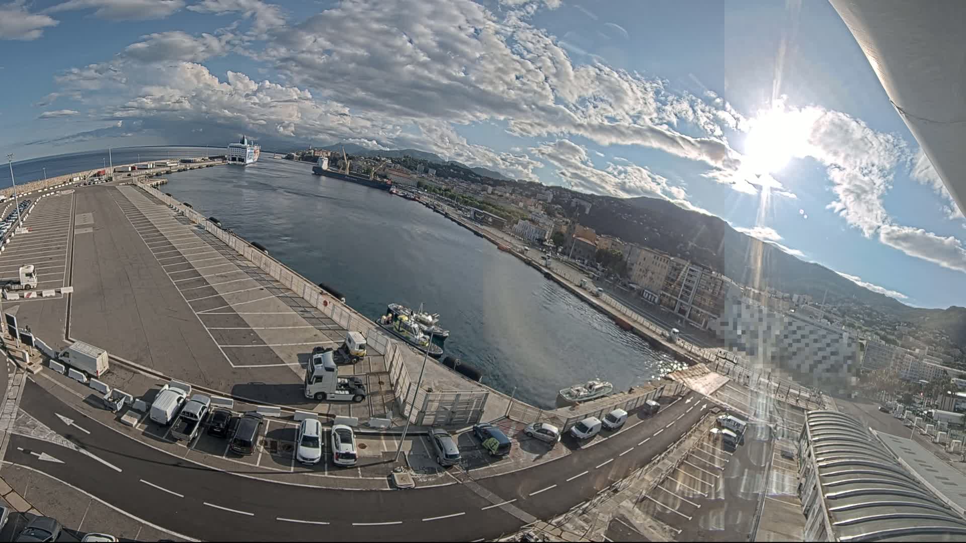 A sunny day reveals a harbor with a large parking lot, several cars, a few small boats, and a ferry in the distance, all nestled against a backdrop of mountains and a city.