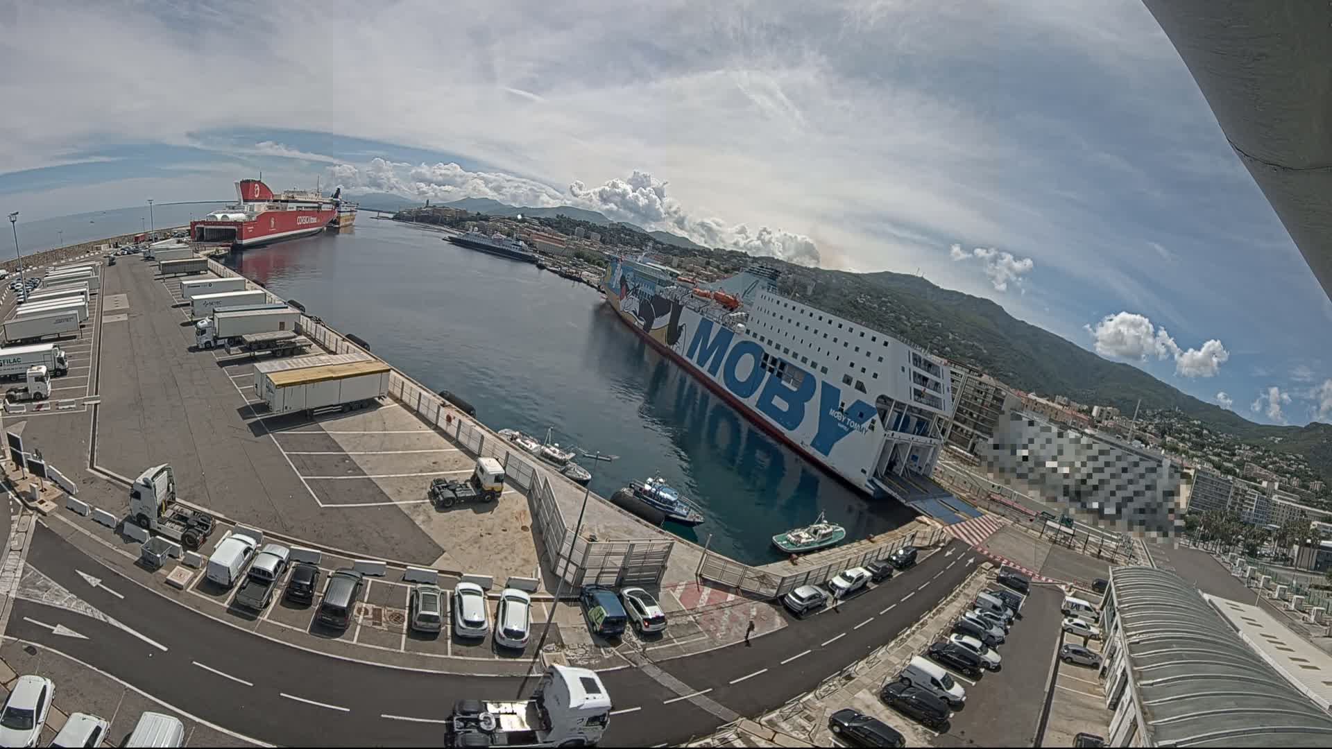 A ferry terminal with two large ferries docked, numerous cars parked in lots, and several trucks and trailers, all under a partly cloudy sky.