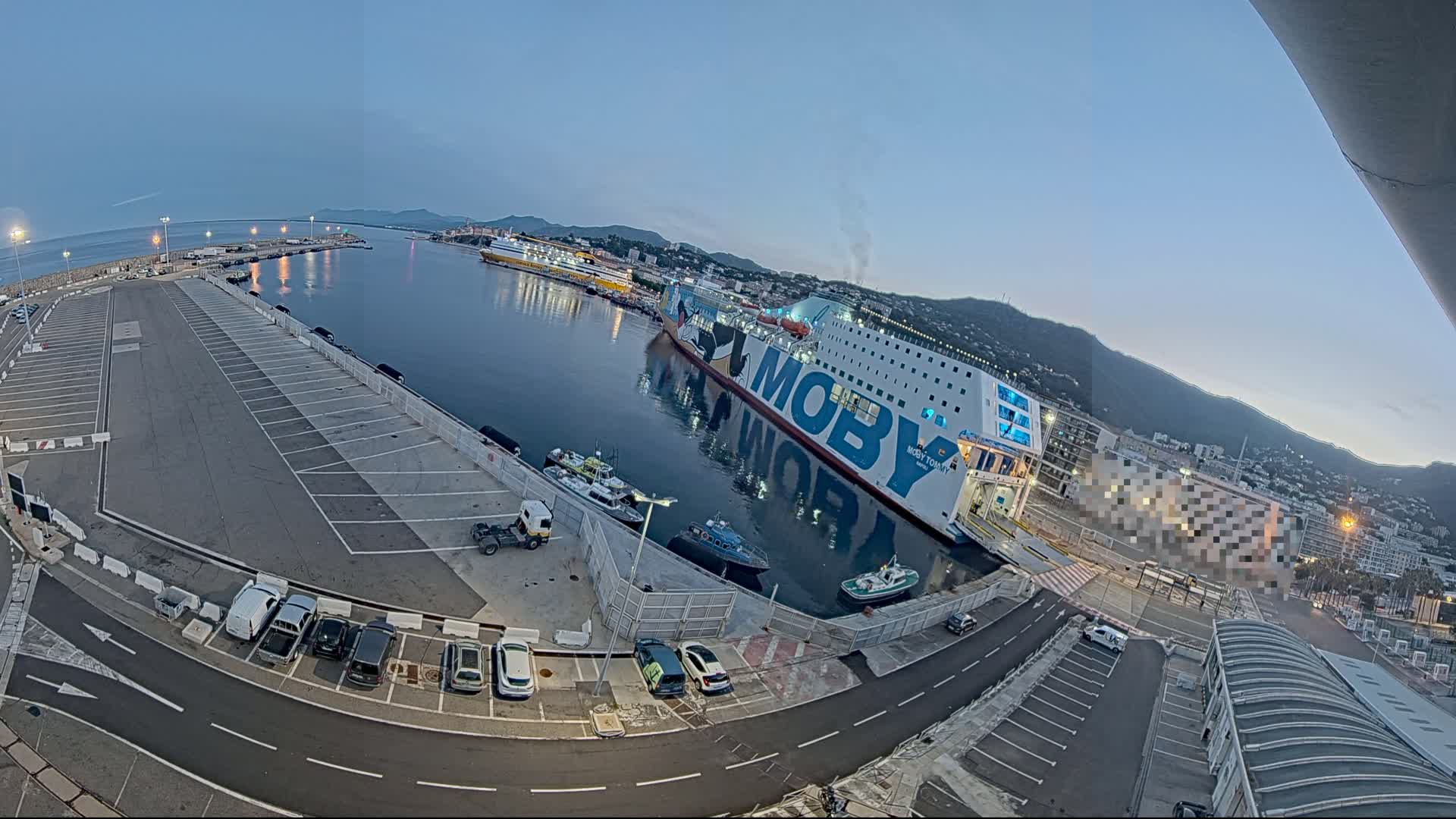 A large ferry is docked at a port under a twilight sky, with several smaller boats and cars visible in a parking lot nearby.
