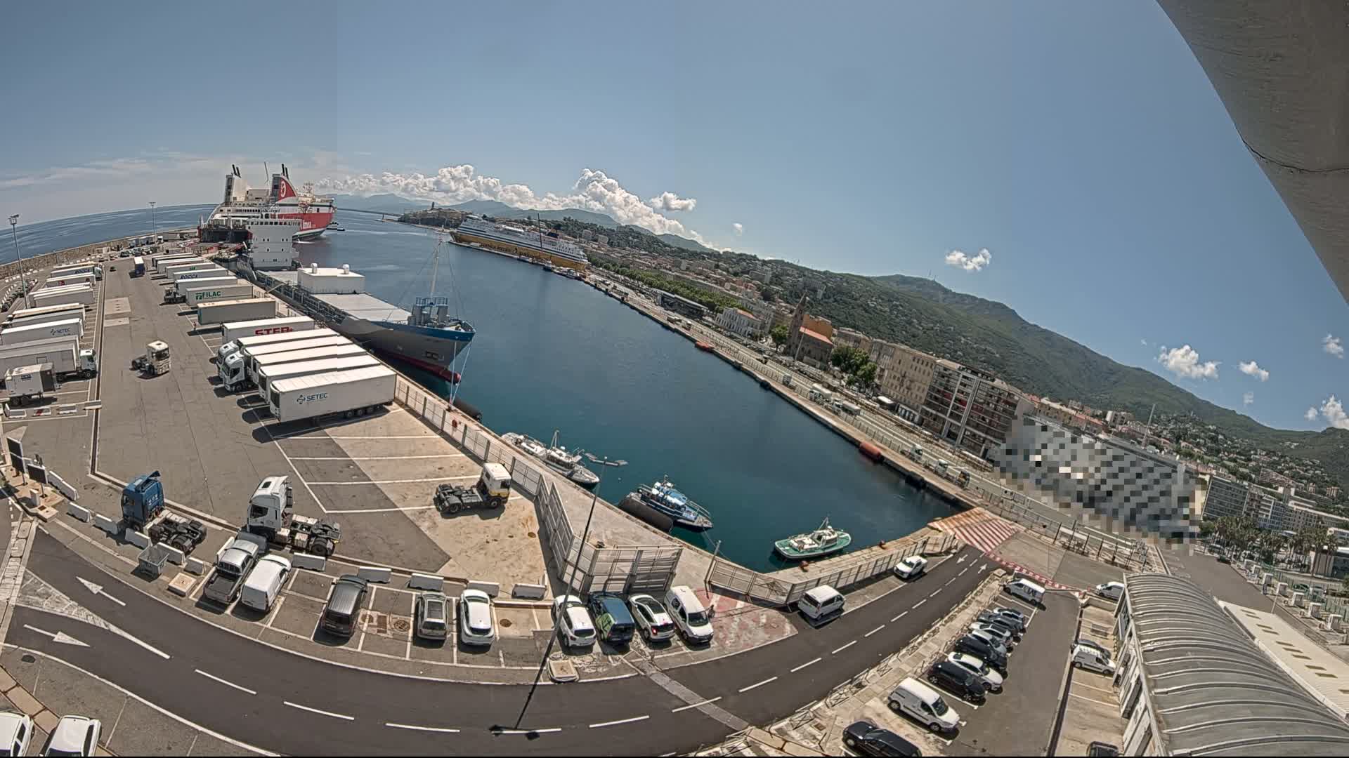 A harbor on a sunny day shows several large ships docked, smaller boats in the water, and numerous cars and trucks in a parking lot next to the docks.
