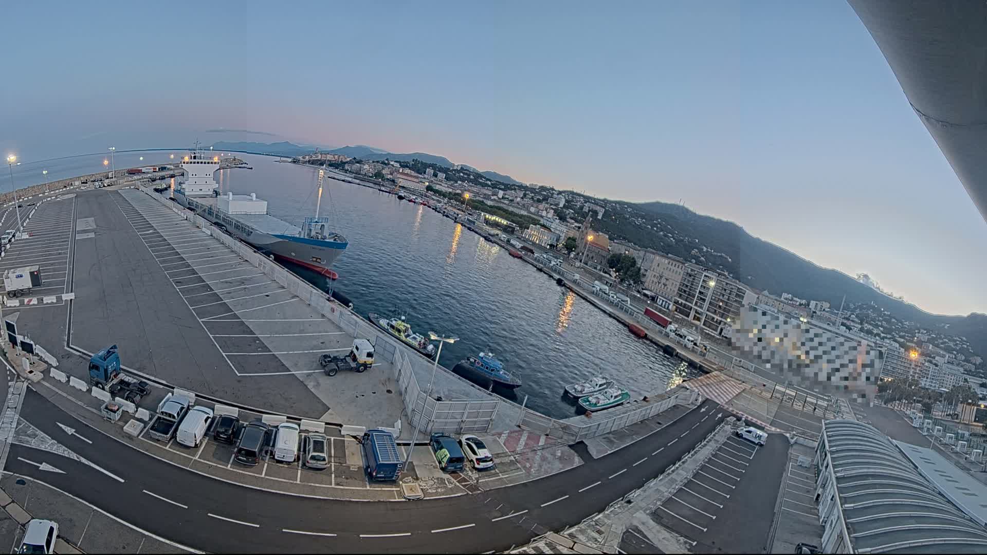 A large cargo ship is docked at a port, with several smaller boats and cars visible in a parking lot under a clear twilight sky.