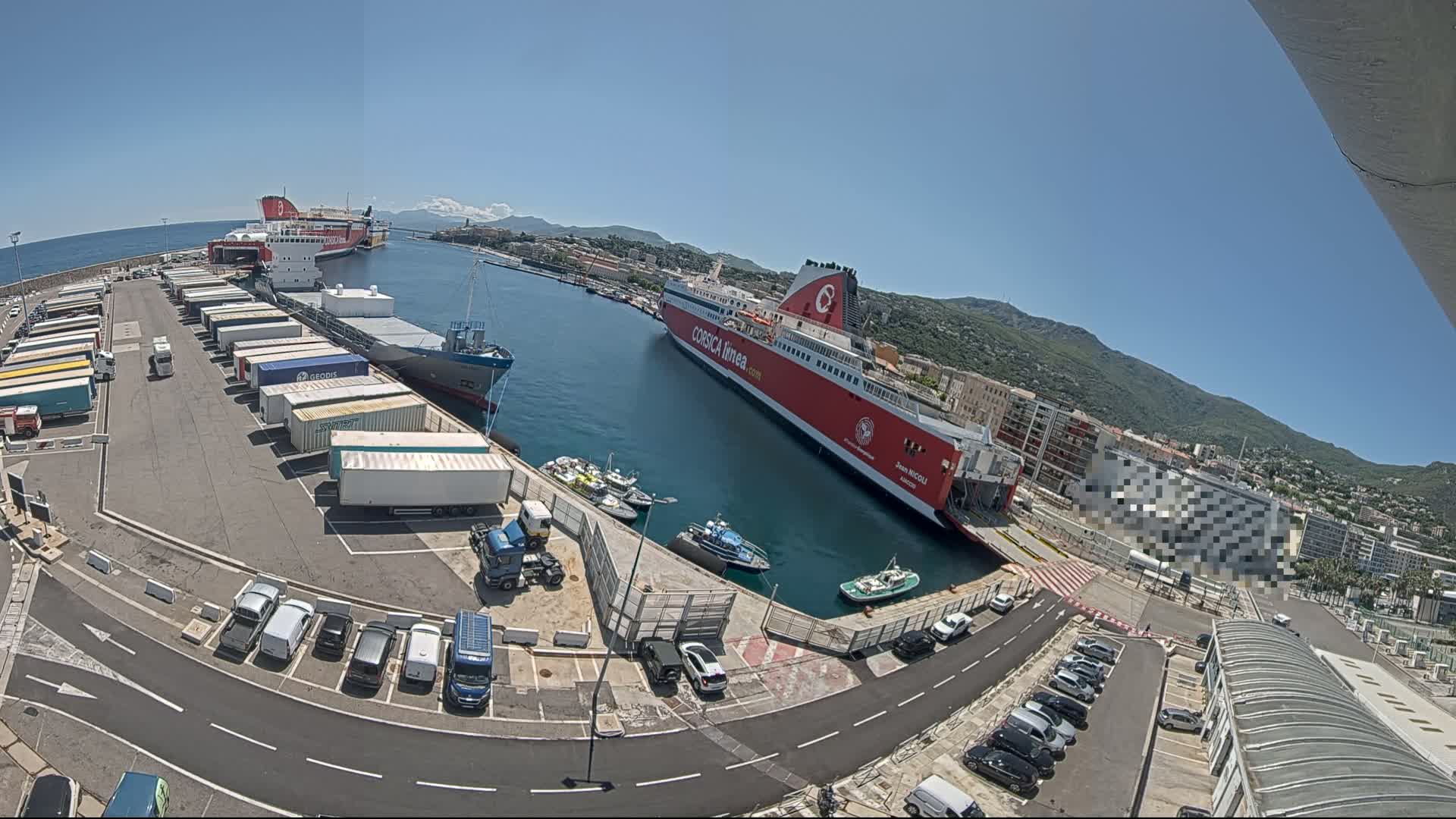 A sunny day at a harbor shows two large ferries docked, surrounded by cargo trucks, smaller boats, and parked cars.