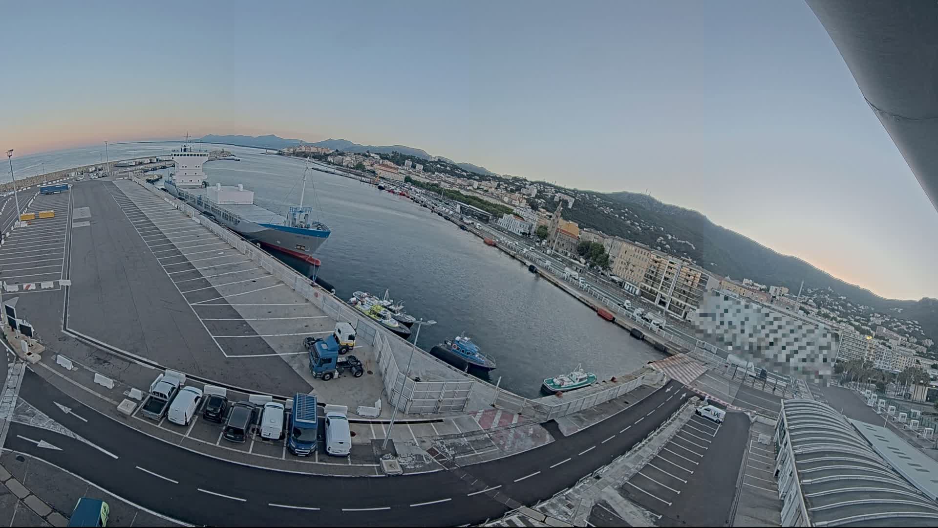 A large cargo ship is docked at a harbor next to a parking lot filled with cars and trucks, under a clear, pale sky; a city and mountains are visible in the background.