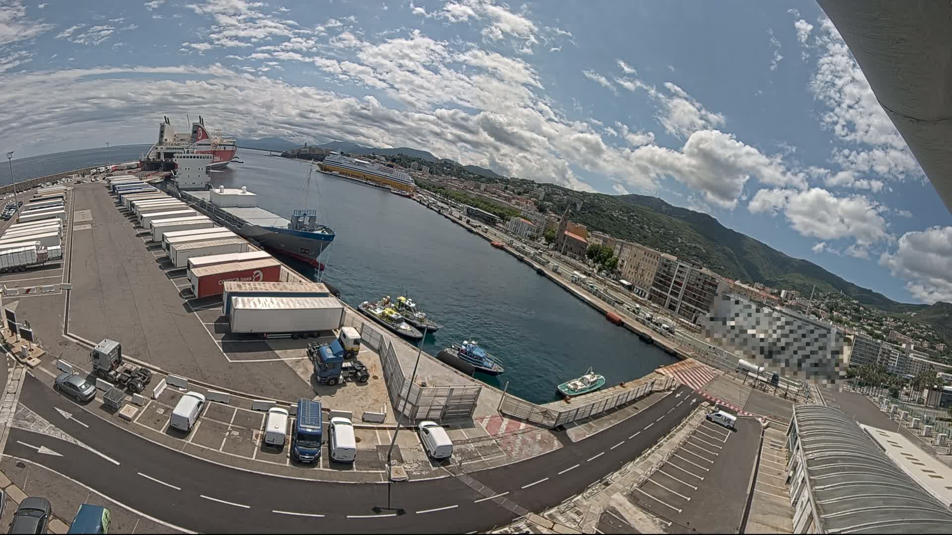 A harbor on a partly sunny day with numerous cargo ships, smaller boats, and trucks parked in a lot next to buildings and mountains in the distance.