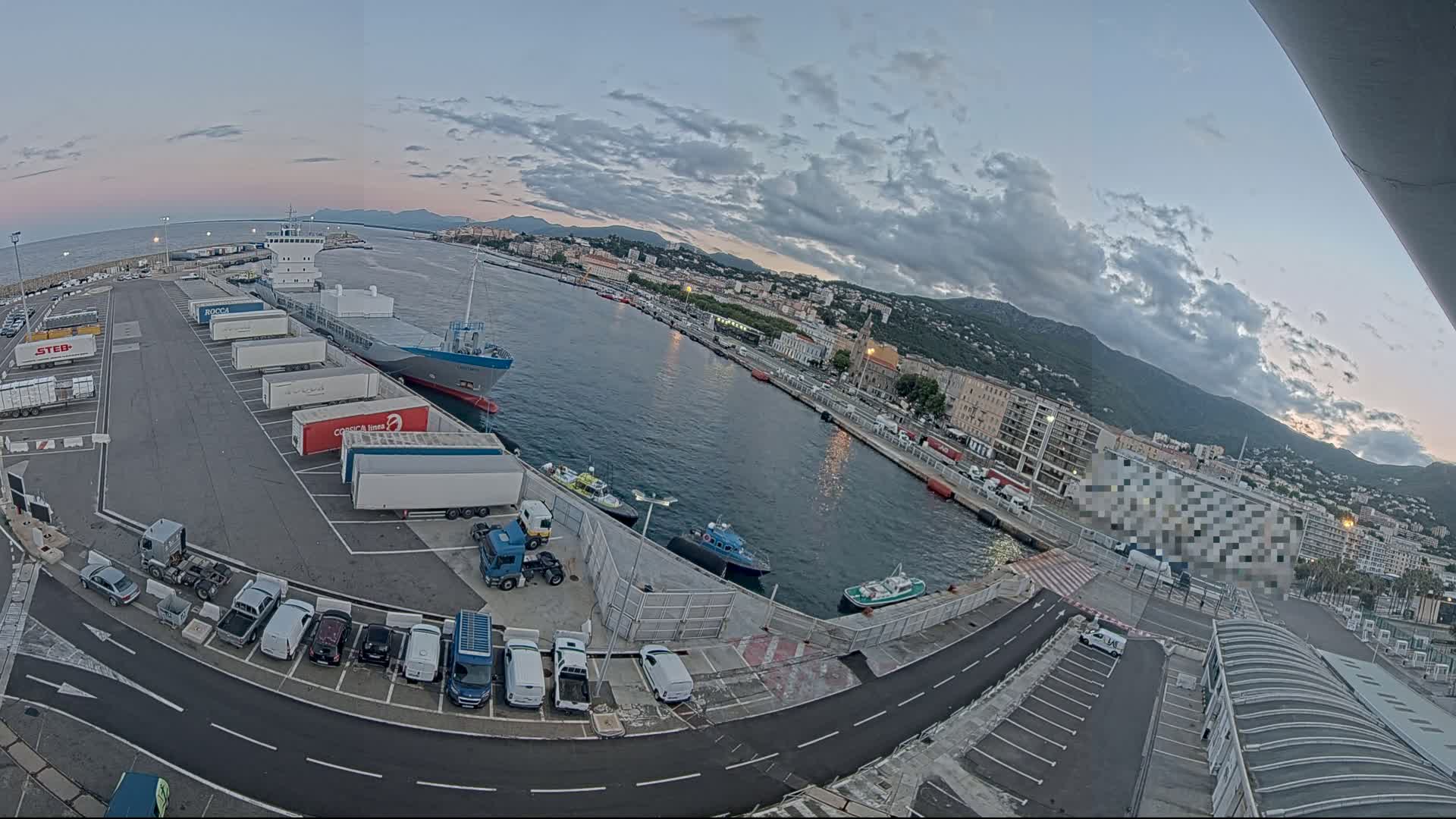A harbor with a large cargo ship and several trucks parked at a dock, under a partly cloudy twilight sky, with a city and mountains in the background.