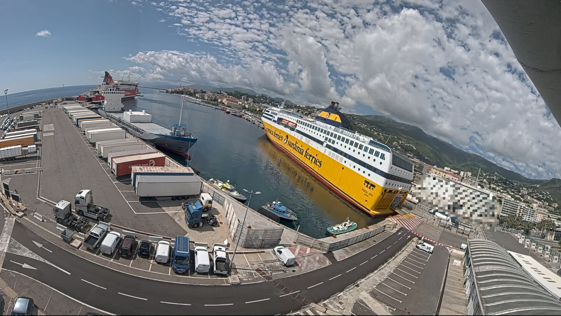 A large yellow ferry is docked at a port on a partly cloudy day, with numerous trucks and cars parked nearby, and other ships in the background.