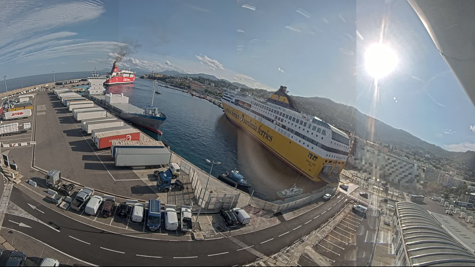 A sunny day at a port shows two large ferries docked, one red and one yellow, with numerous trucks and cars parked in the surrounding area.