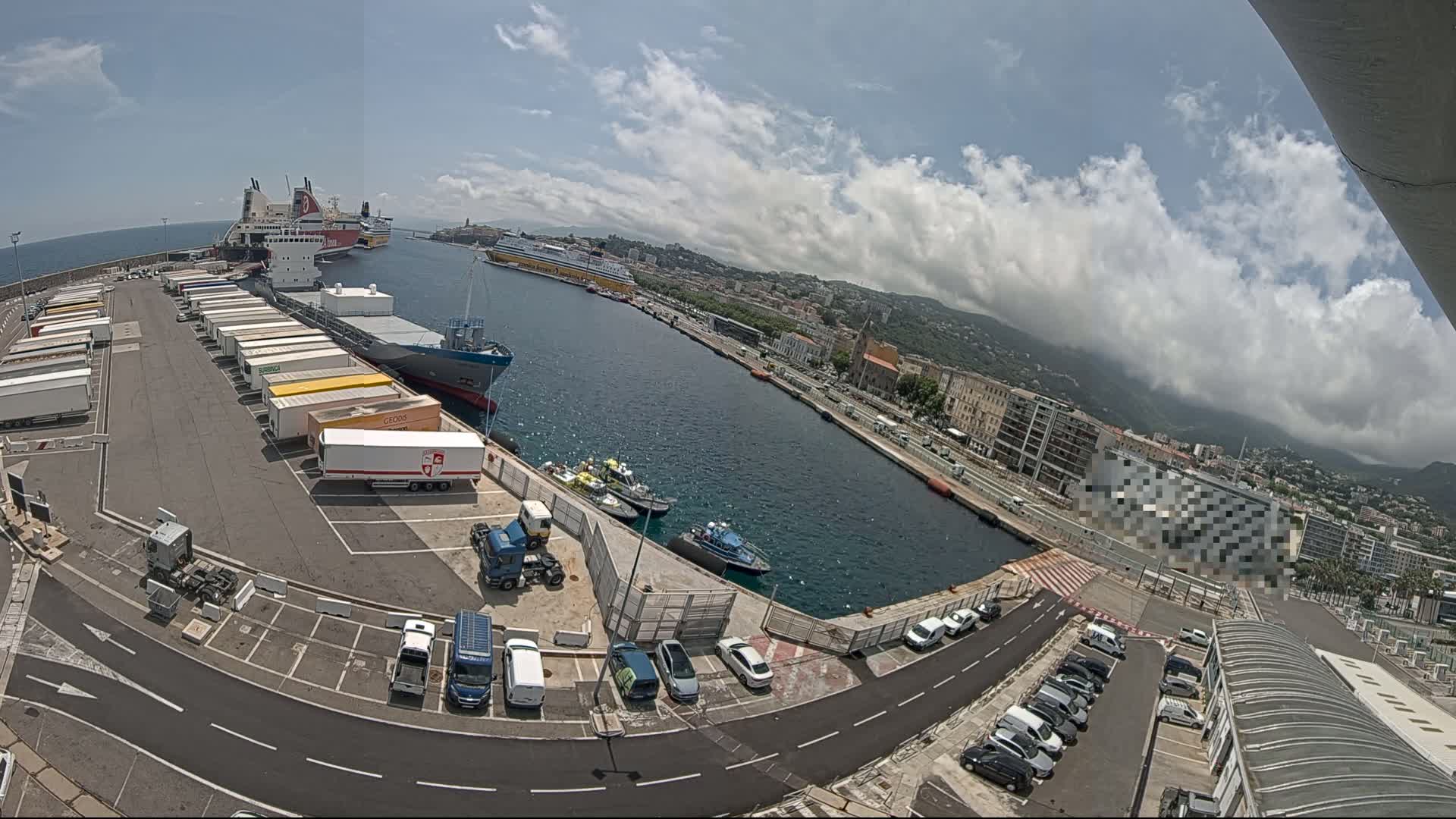 A sunny day at a harbor shows several large ships docked, numerous cargo trucks parked nearby, smaller boats in the water, and cars parked in a lot overlooking the water and a city in the distance.