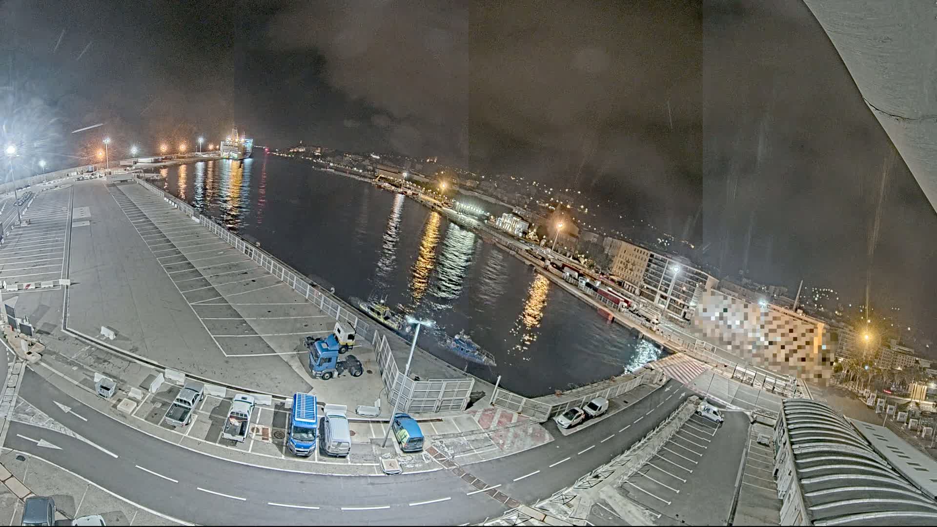 A nighttime, slightly rainy view of a harbor with a large empty parking lot, several small vehicles, and a few boats in the water.