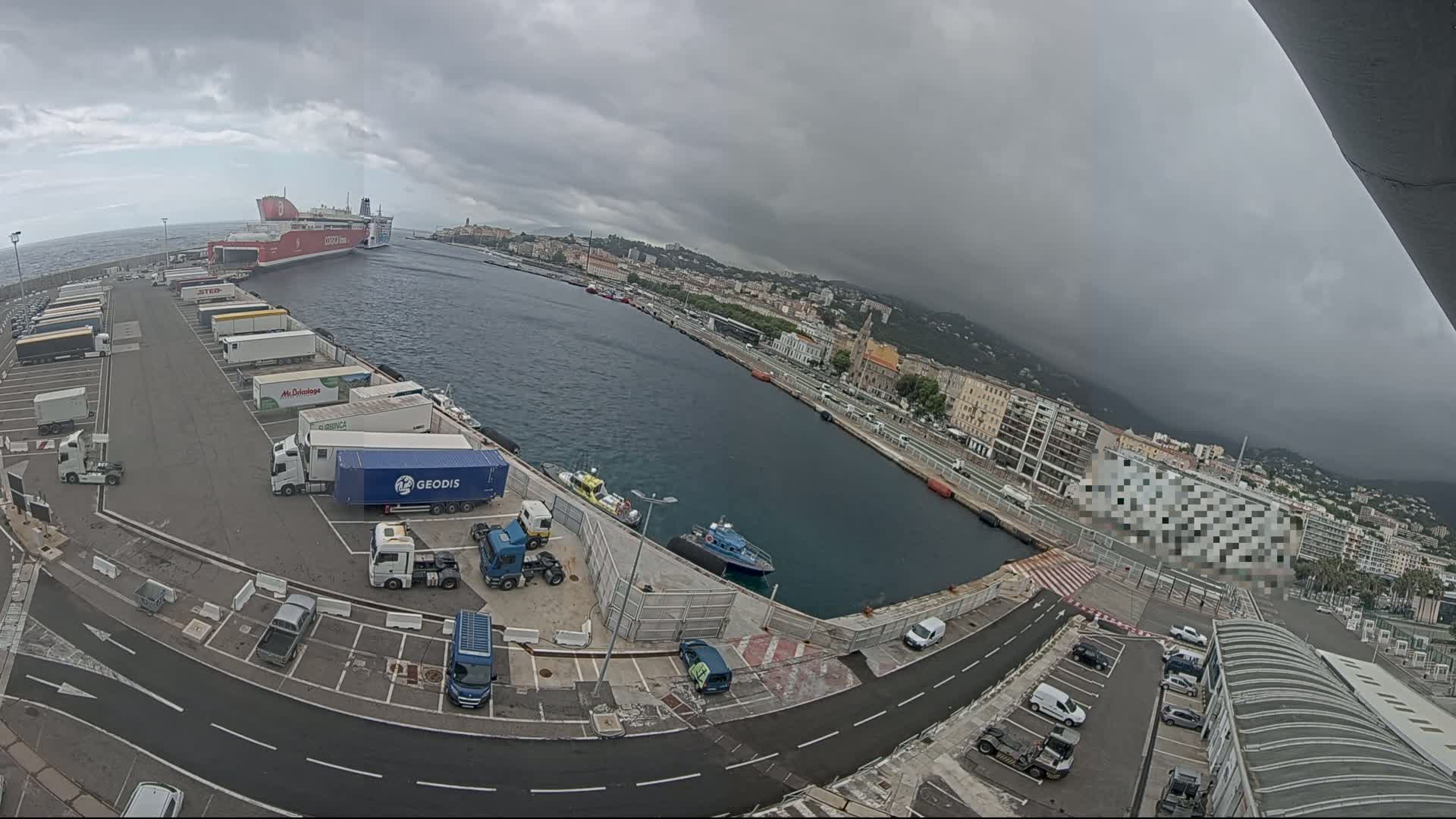 A ferry is docked at a harbor under an overcast sky, with numerous trucks parked in the adjacent lot and smaller vessels in the water.