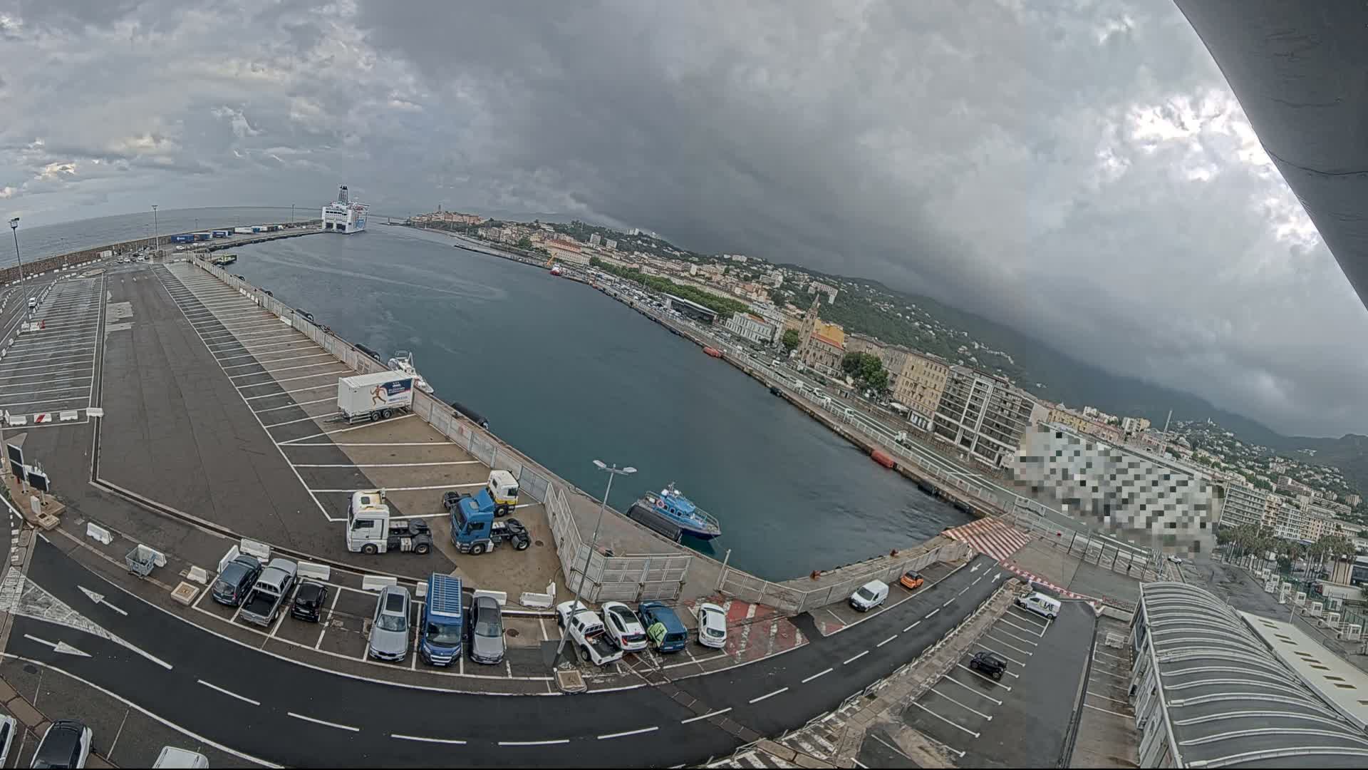 A high-angle view of a harbor on an overcast day shows a ferry docked, cars parked in a lot, trucks, and a small boat in the water, all under a cloudy sky.