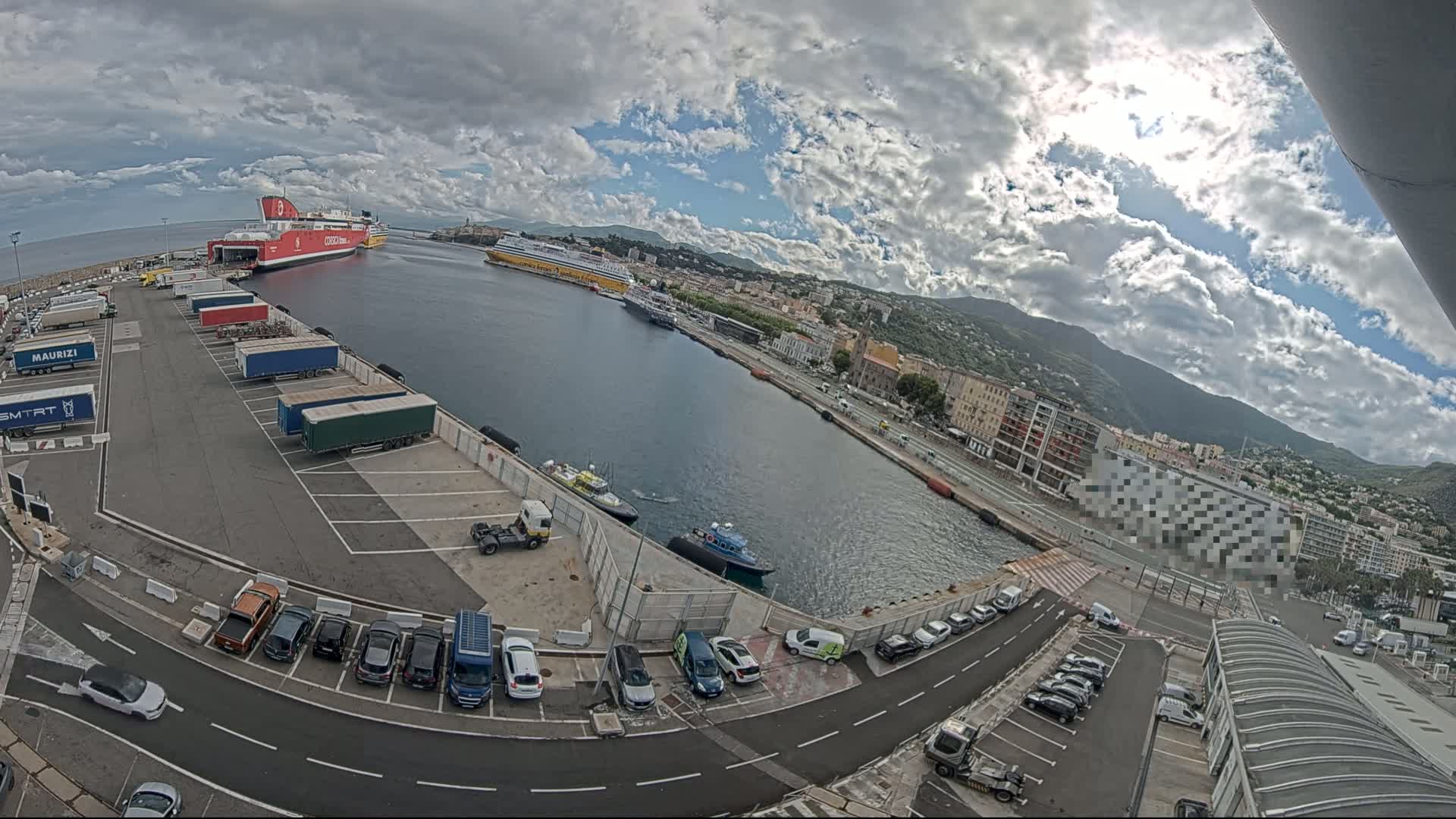 A partly cloudy day at a harbor shows two large ferries docked, numerous cars parked in lots, and several trucks and smaller vehicles near a waterfront city nestled against mountains.