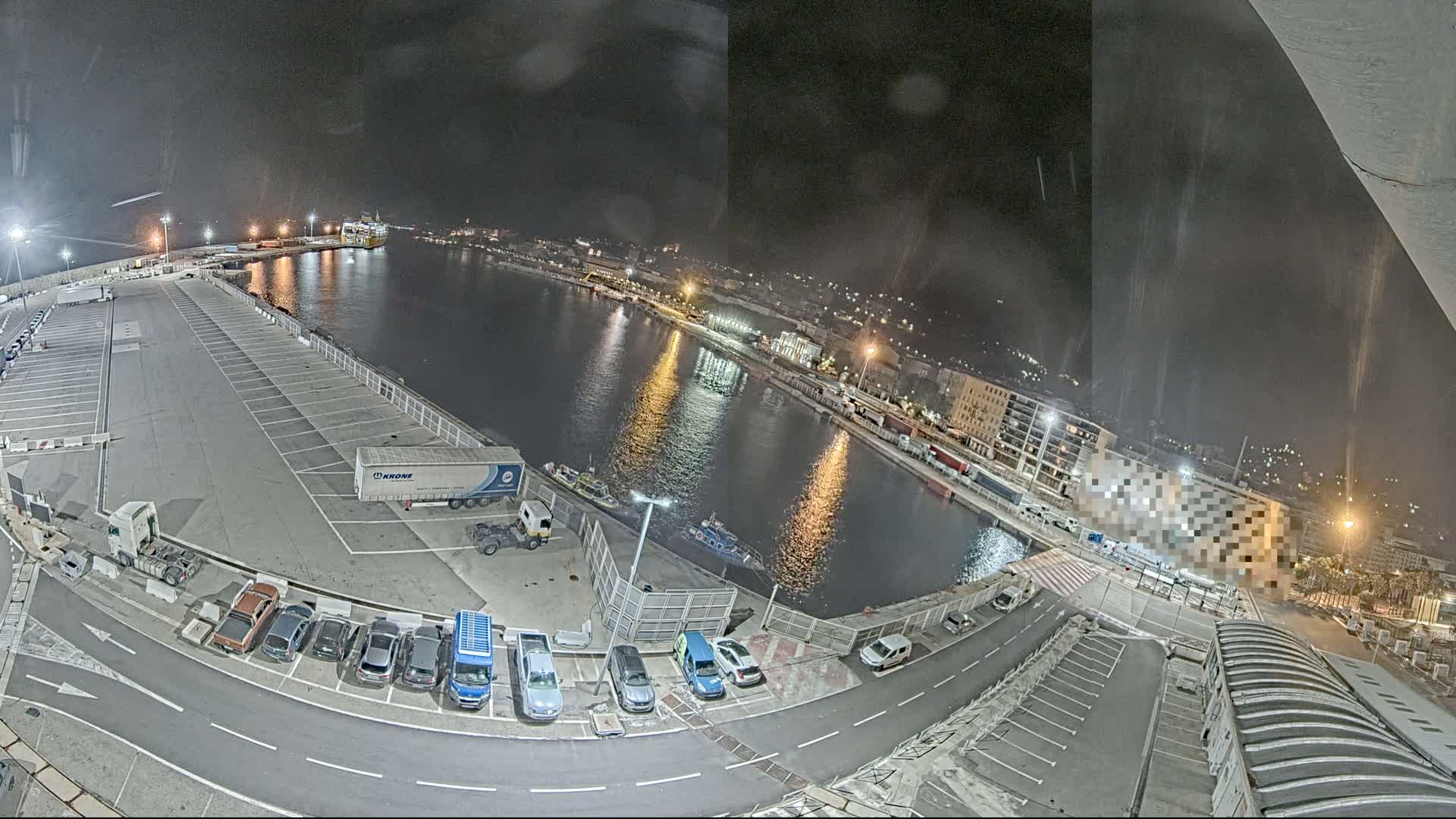 A nighttime, high-angle view of a harbor with a large empty parking lot, several cars, and a few trucks near a calm body of water reflecting city lights.