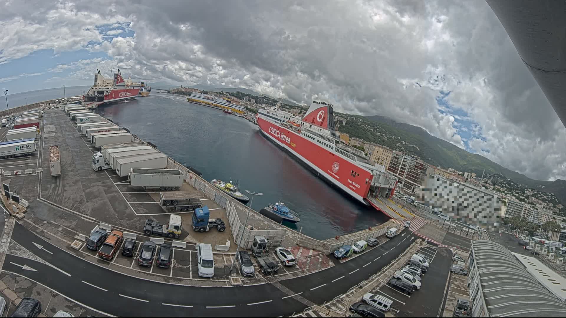 A large red ferry is docked at a port, with several other ships and numerous trucks and cars in the parking lot under cloudy skies.