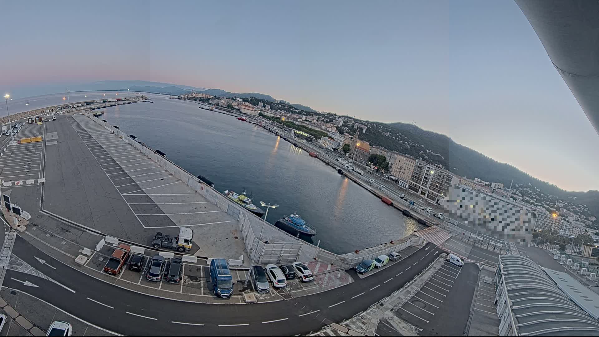 A large empty parking lot overlooks a calm harbor with several small boats and a city nestled against a mountain range under a clear twilight sky.